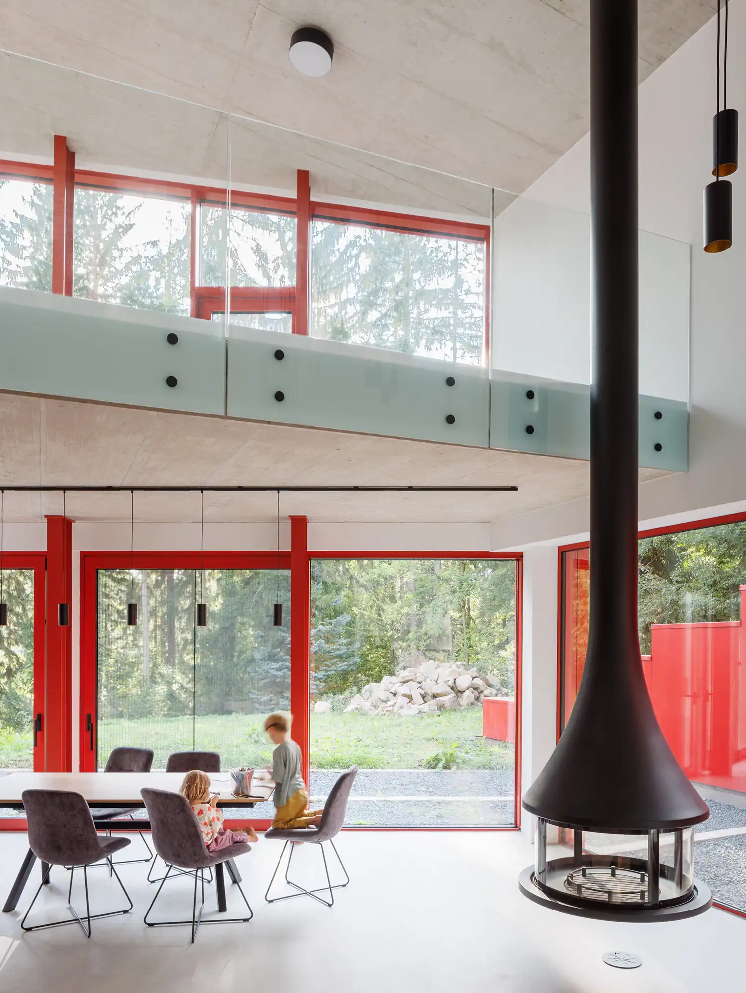 Interior living space with double-height glass walls, red window frames, a black hanging fireplace, and raw concrete ceilings.