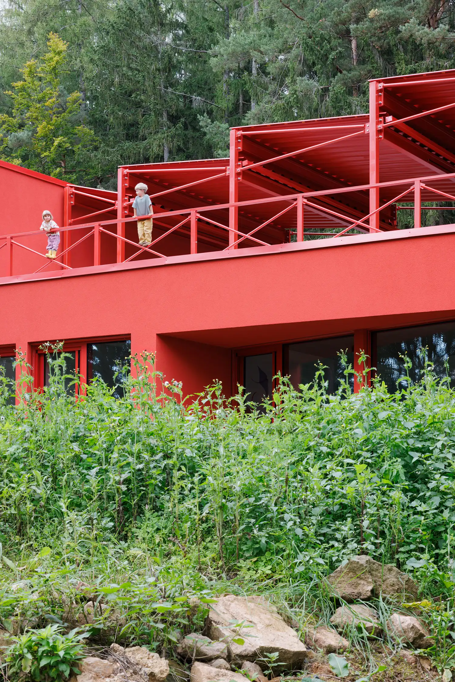 Low-angle view of children standing on a red balcony under a sawtooth roof, surrounded by wild garden plants.