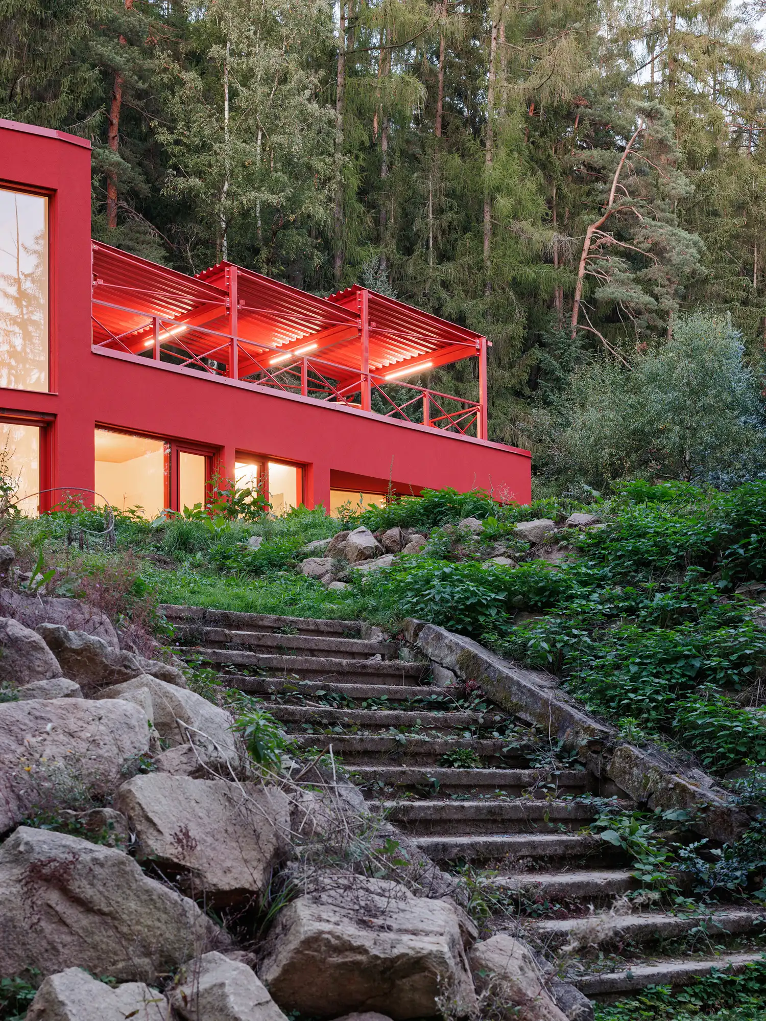 Evening shot of the red villa with illuminated interior lights and old stone steps leading up through a wild garden.