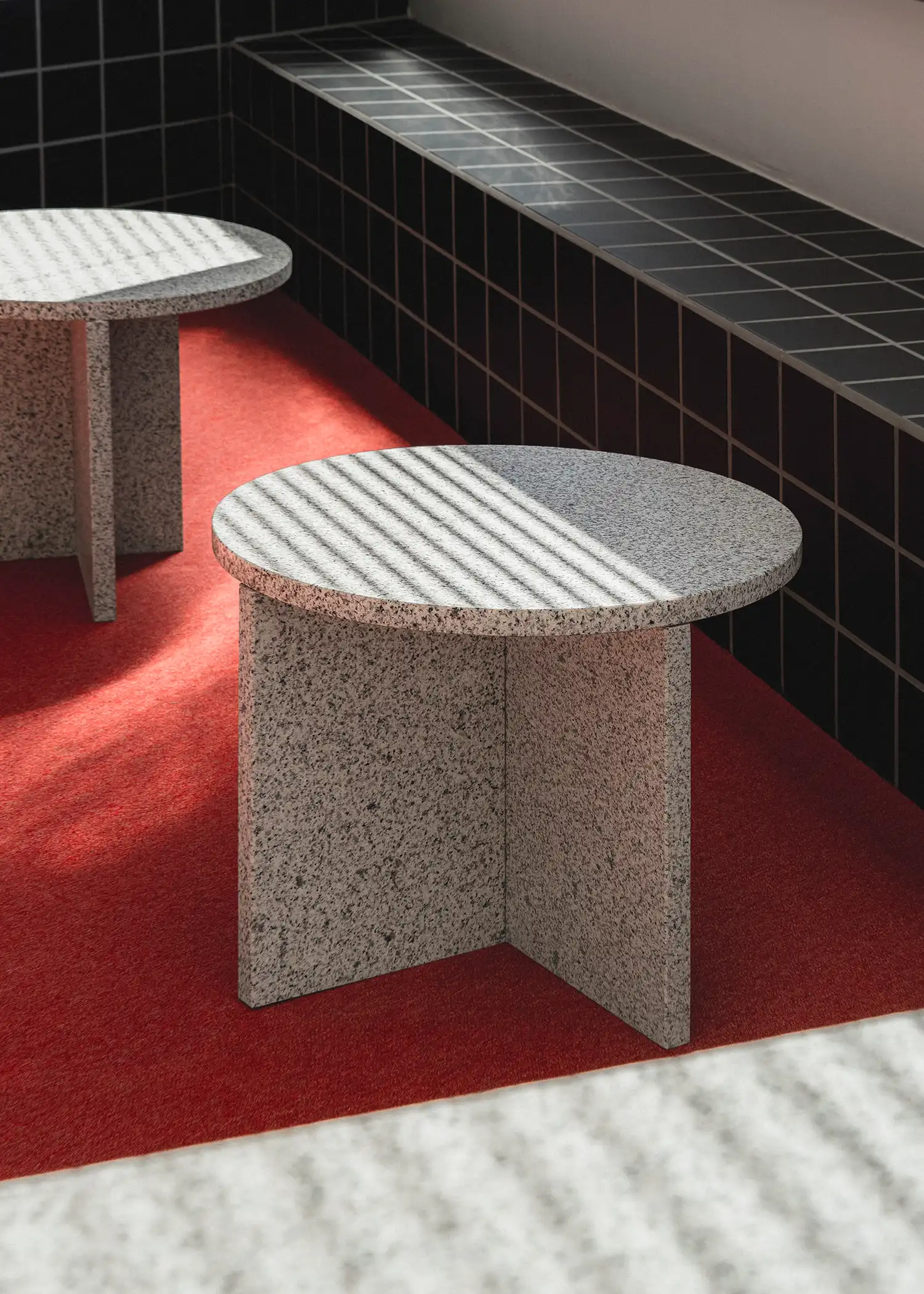 Two minimalist circular granite side tables on a red carpet with striped sunlight shadows.
