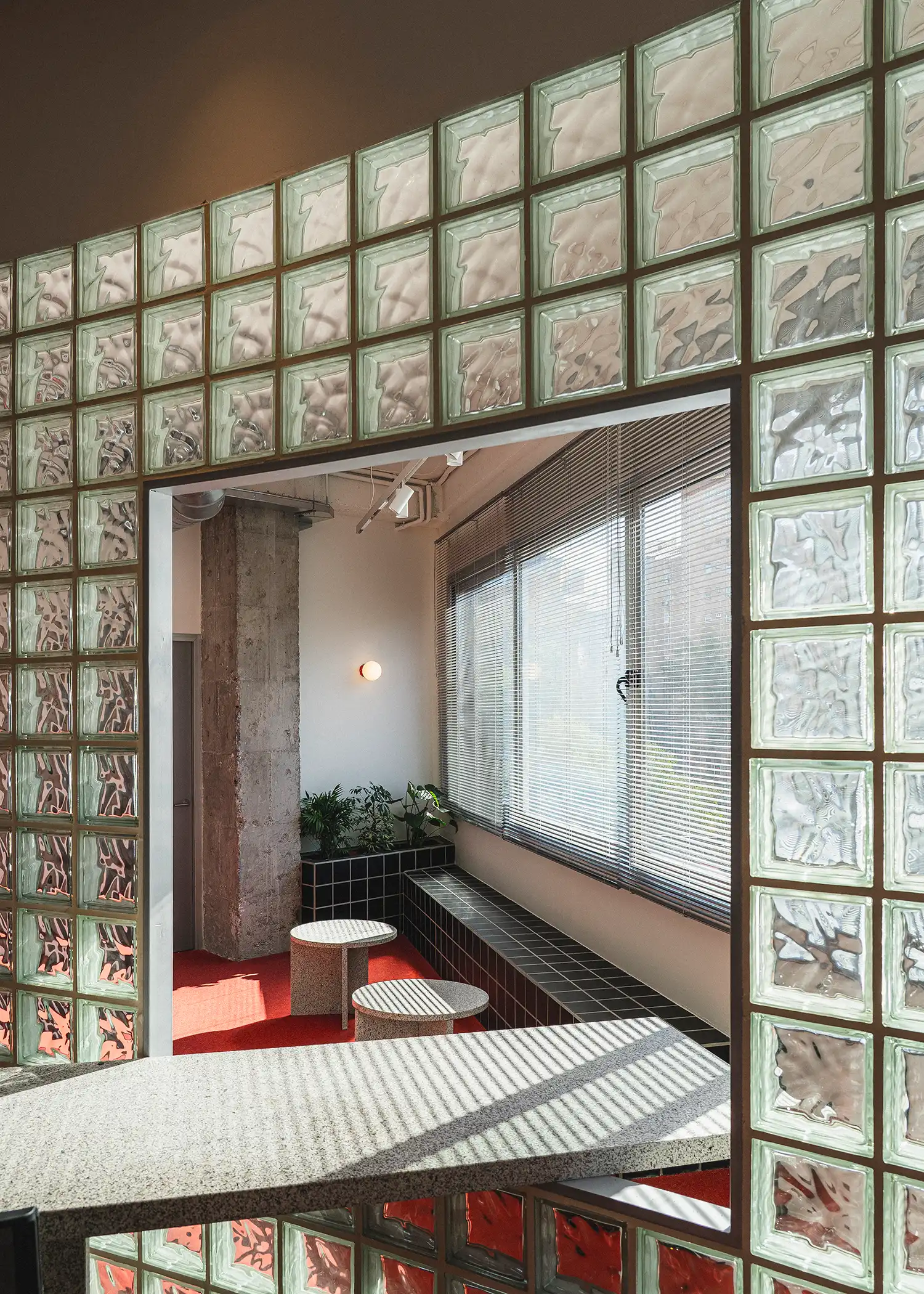 A view through a rectangular opening in a glass-block wall toward a window with blinds.