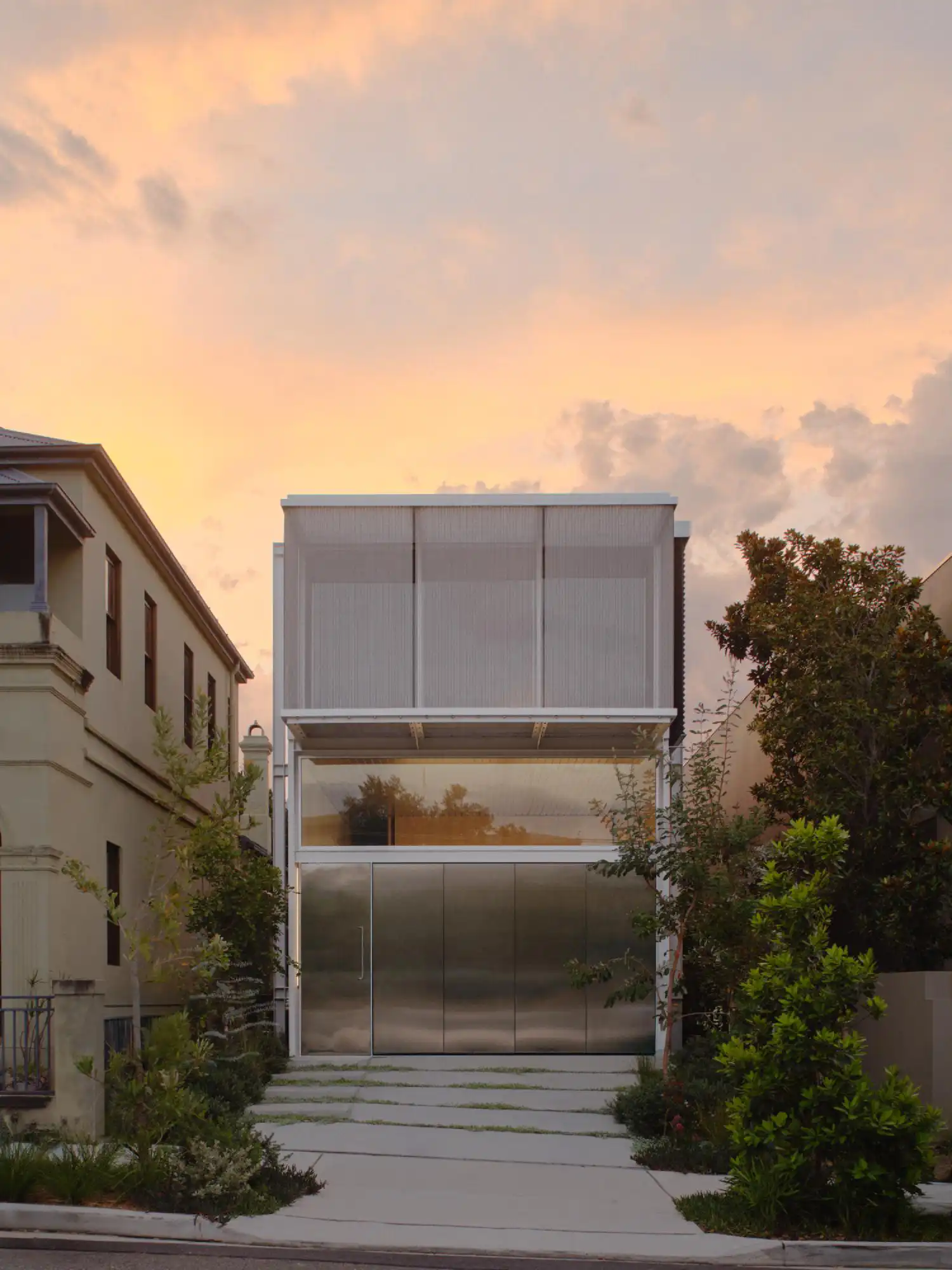 Two-story modern house with a metallic ground floor and a screened upper terrace at sunset.