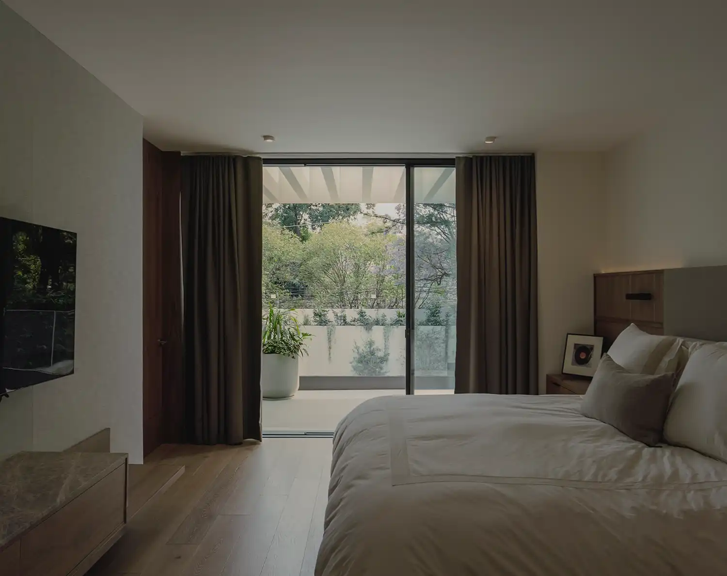 Minimalist master bedroom with neutral bedding and a balcony overlooking green trees.