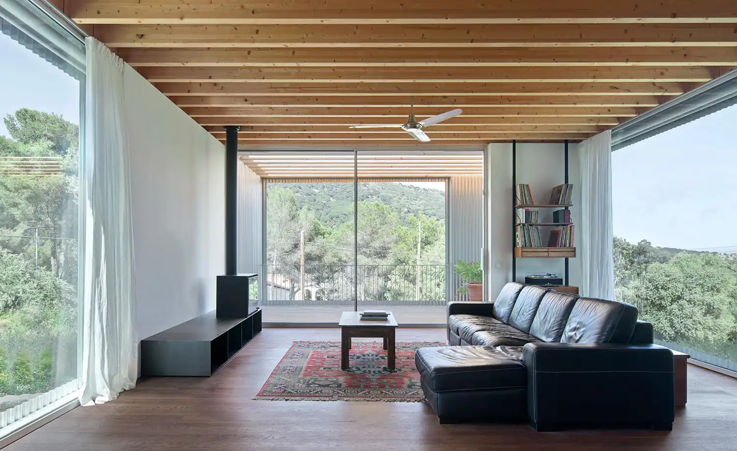 Minimalist living room with exposed timber ceiling beams, a black leather sofa, and panoramic windows.