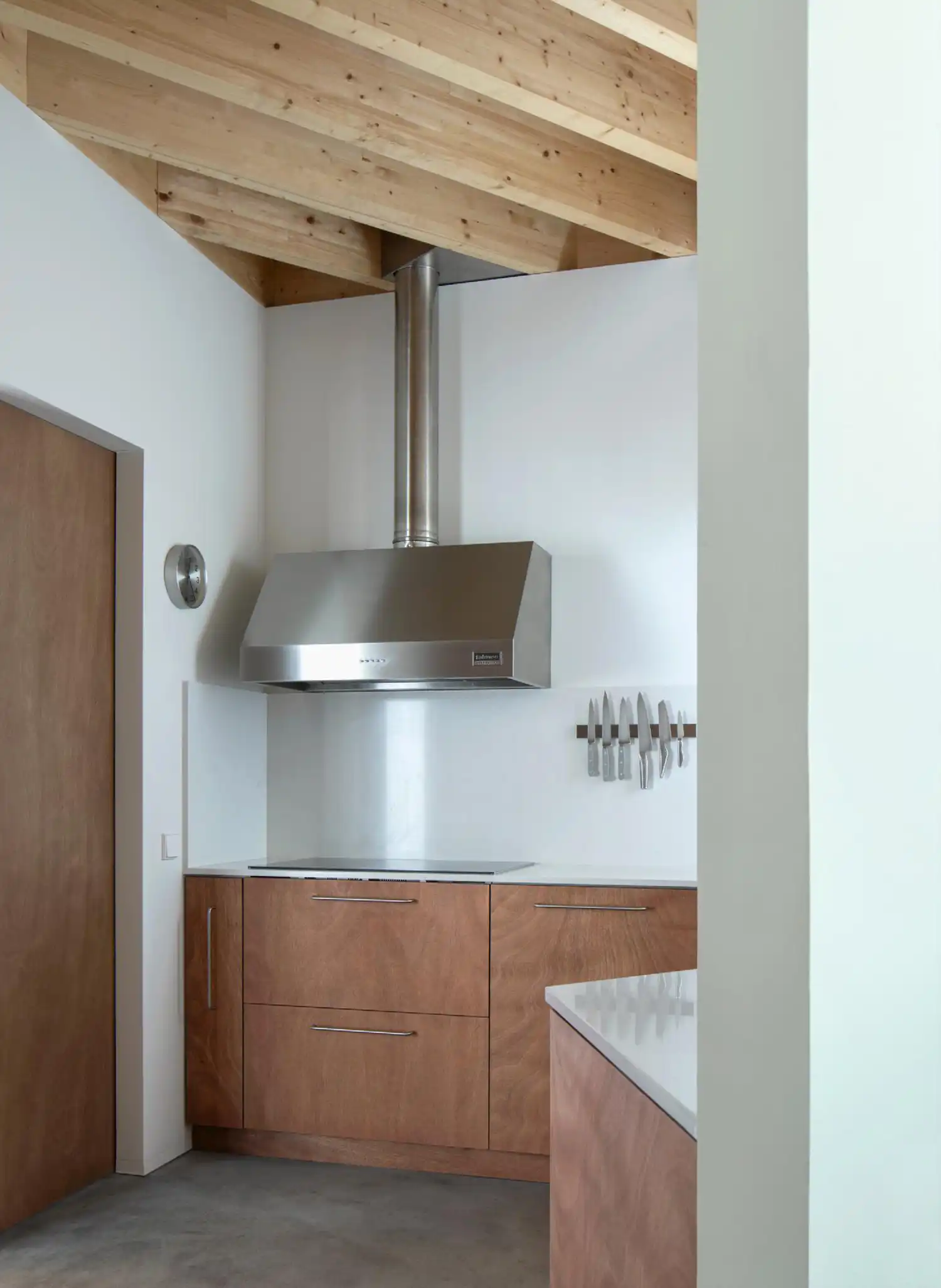 Modern kitchen corner with wooden cabinetry, white countertops, and a stainless steel vent hood under timber beams.