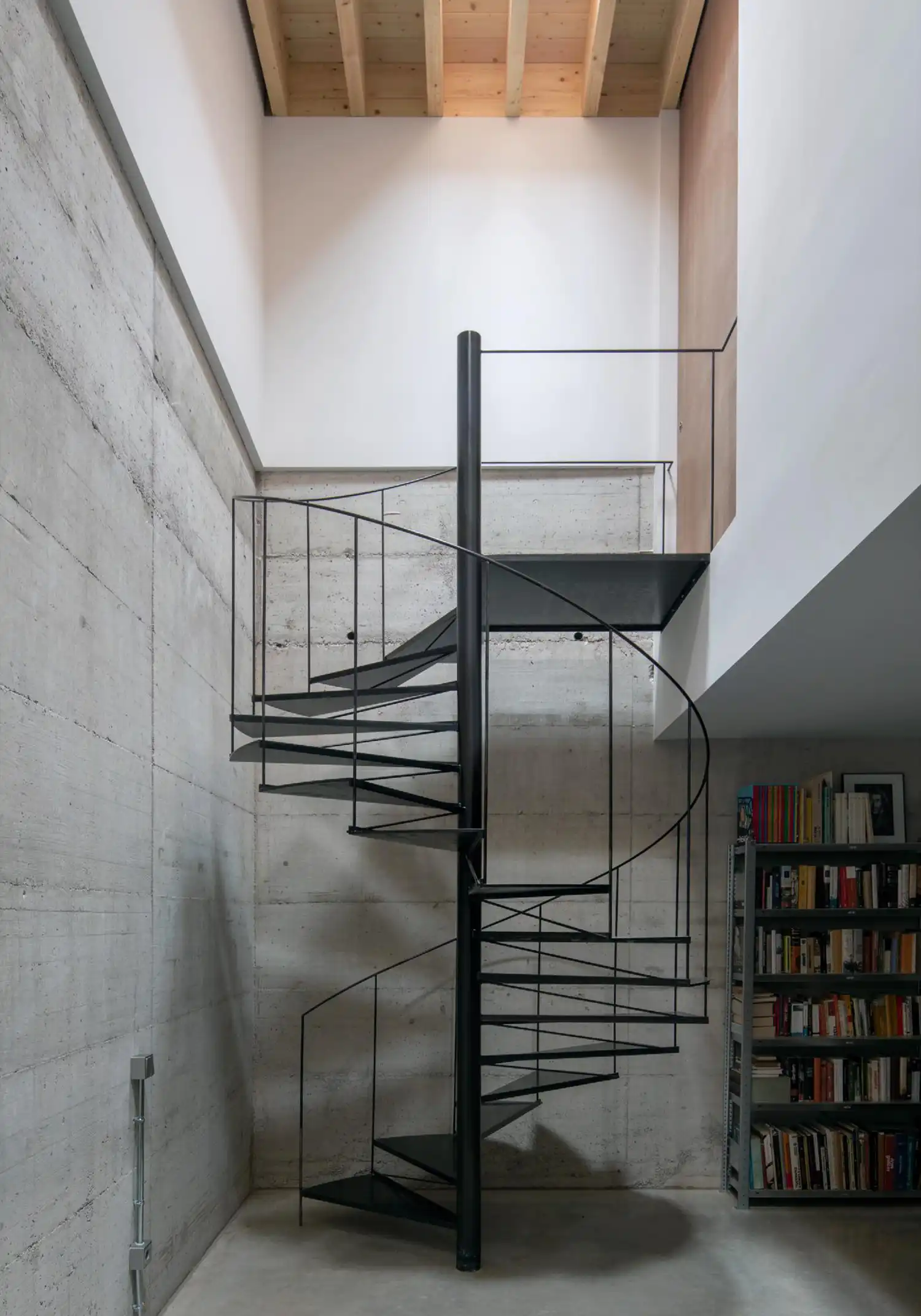 A black metal spiral staircase in a concrete-walled room leading up to a timber ceiling.