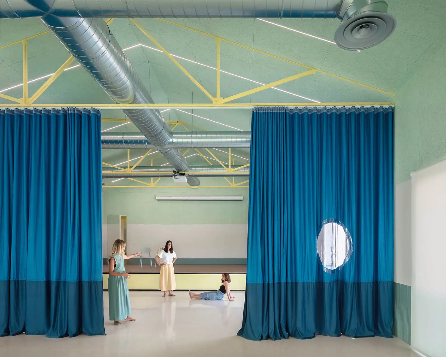 Residents interacting in the multipurpose hall divided by teal acoustic curtains and industrial ceiling details.