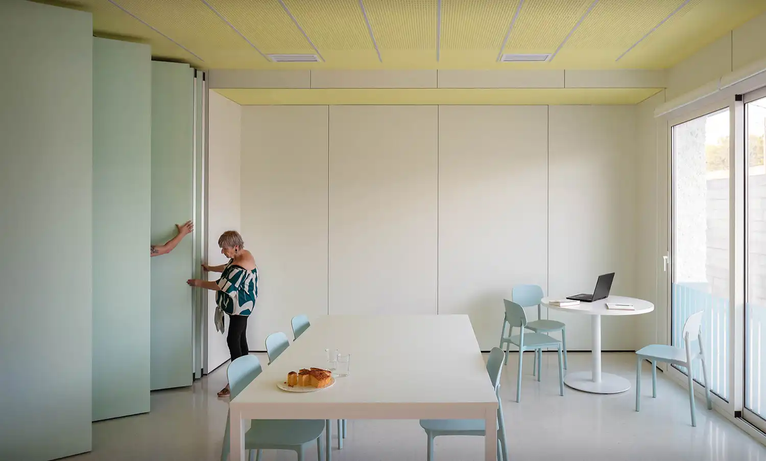 A person moving a sliding partition wall in a bright, minimalist multipurpose room with light blue chairs.