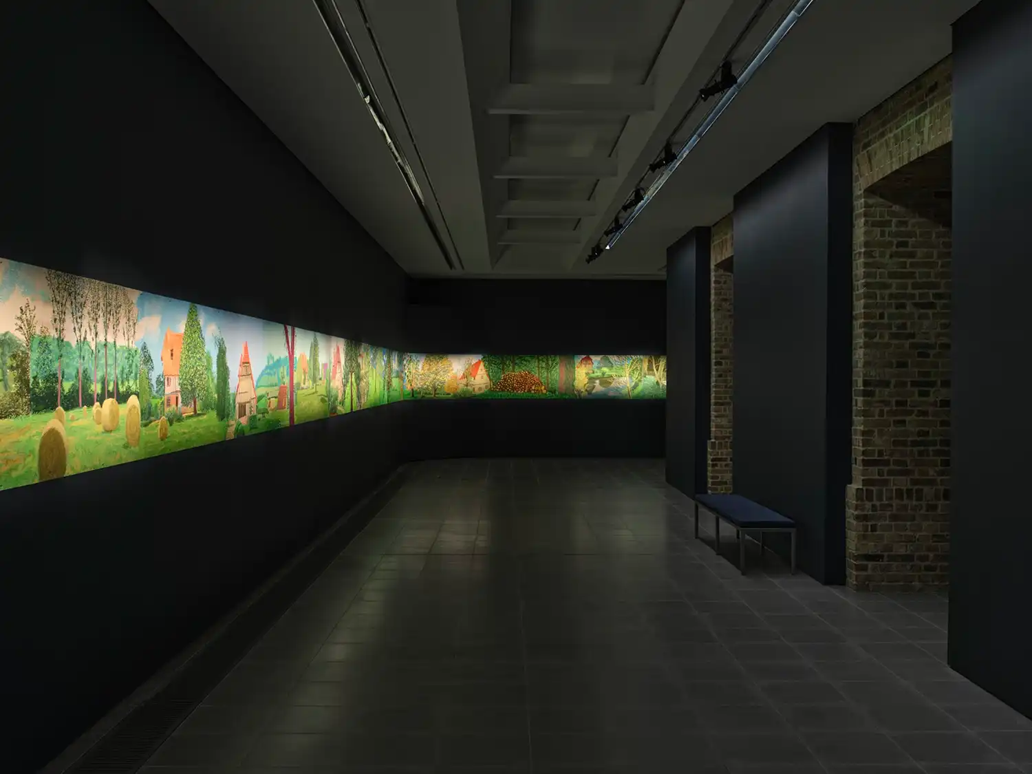 Perspective view down a dark gallery corridor with David Hockney’s continuous digital frieze stretching along the left wall.
