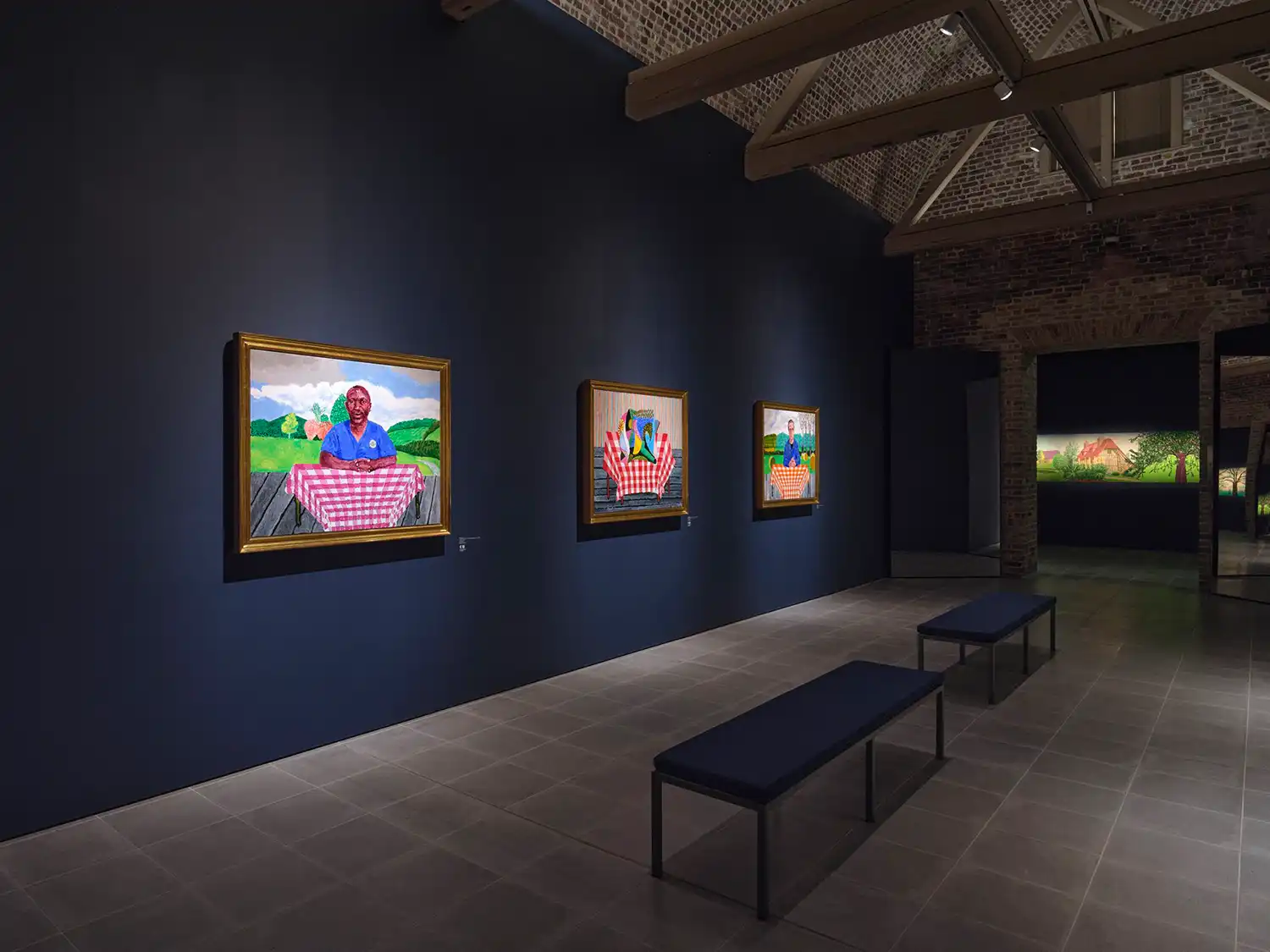 Three framed David Hockney portraits on a dark blue wall in a gallery room with exposed brick arches.