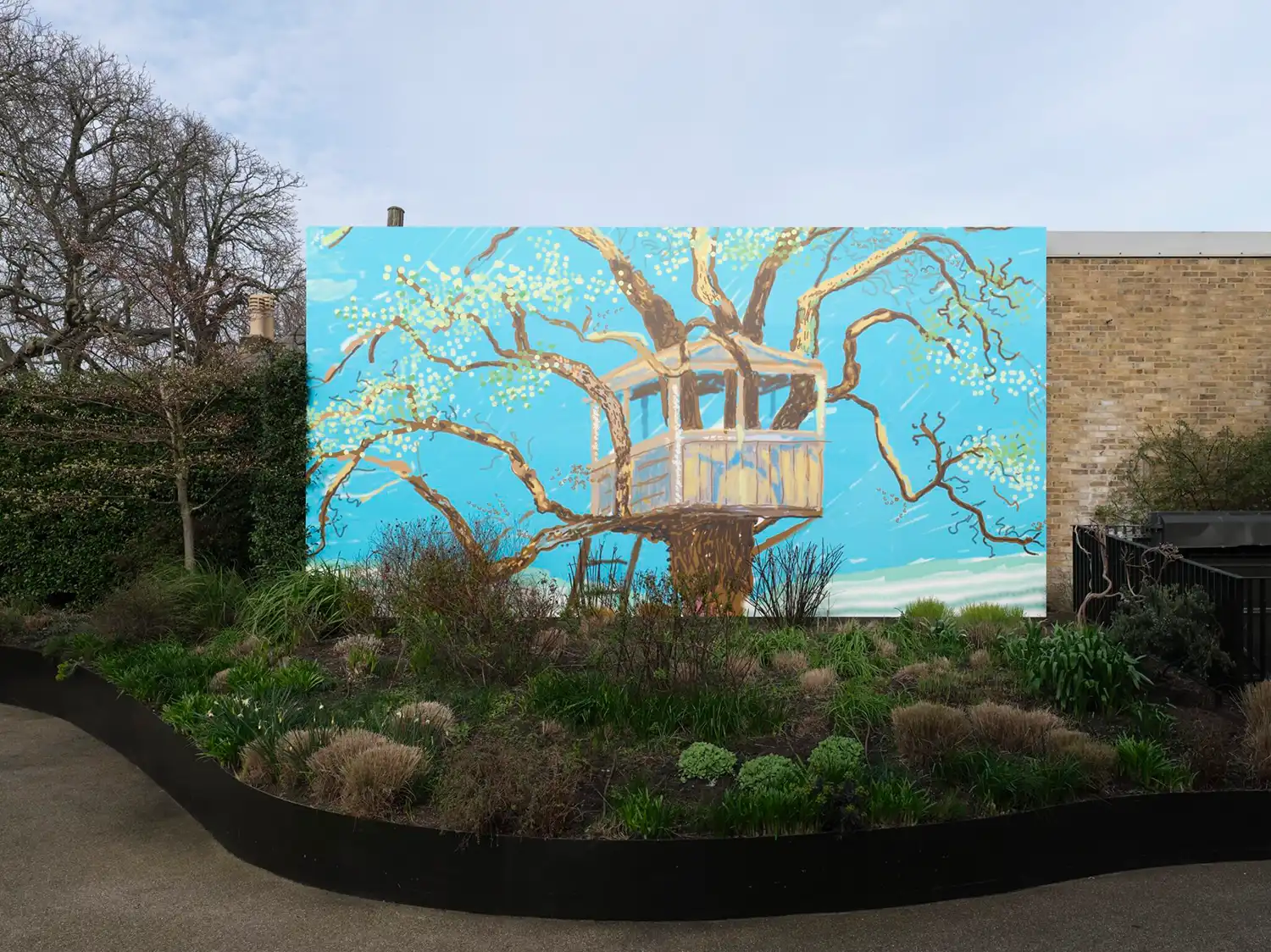 A large rectangular mural of a treehouse in a garden setting, positioned against a brick exterior wall.