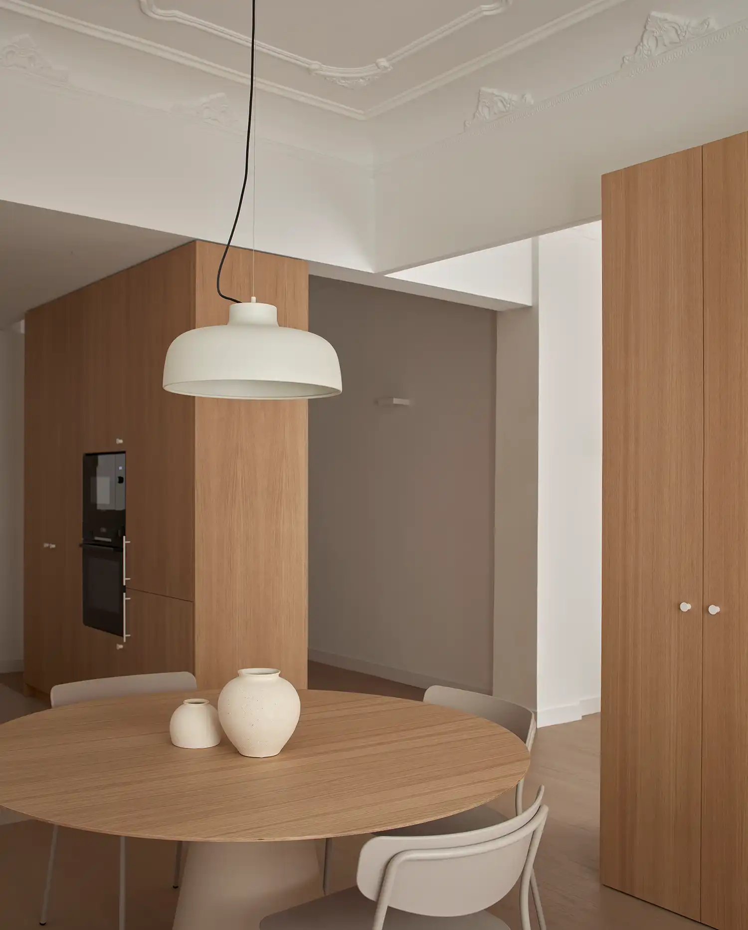 Minimalist dining area with round wooden table and white pendant lamp beneath historic moldings.