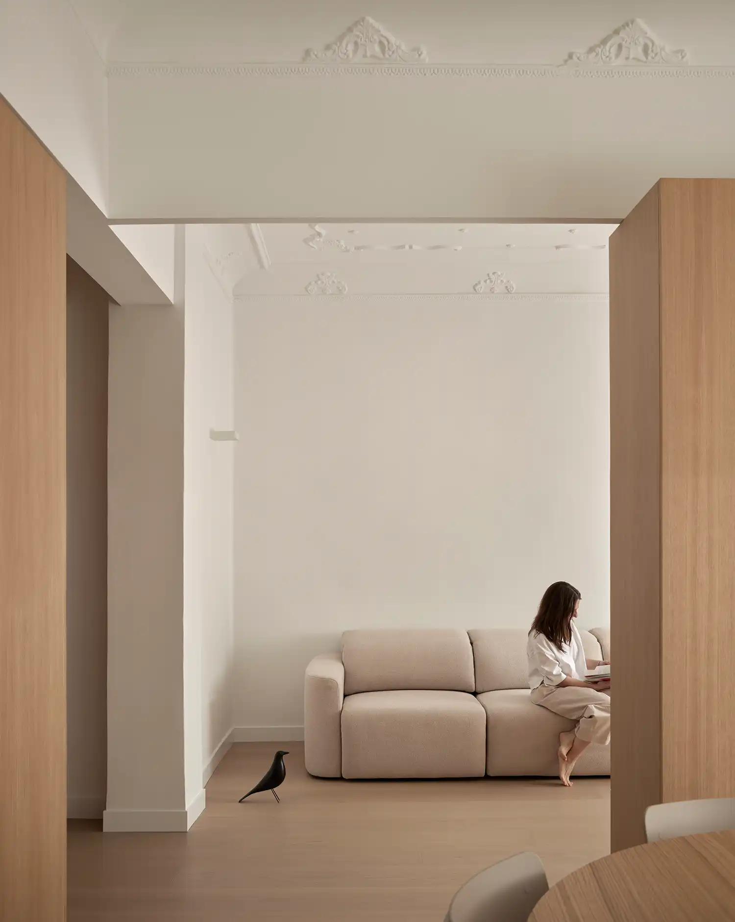 Woman sitting on a neutral sofa in a minimalist living room with white walls and ornate ceiling.