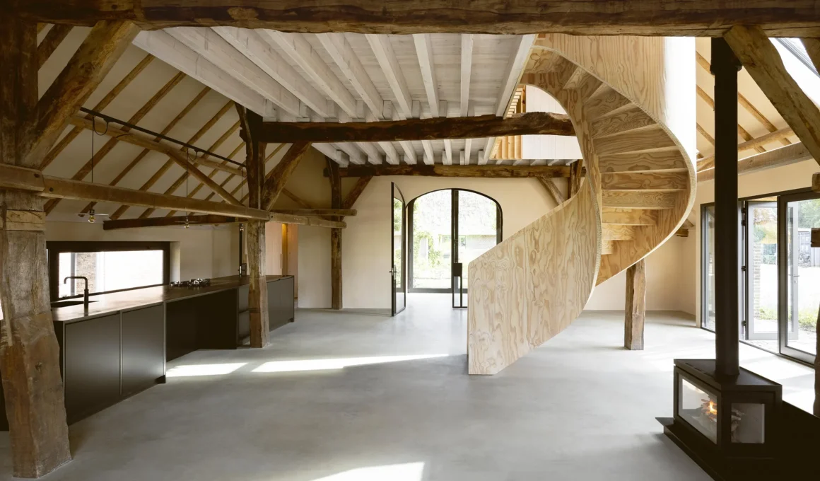 Open-plan barn interior with a helical plywood staircase, concrete floors, and exposed timber beams.