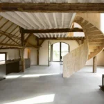 Open-plan barn interior with a helical plywood staircase, concrete floors, and exposed timber beams.