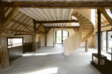 Open-plan barn interior with a helical plywood staircase, concrete floors, and exposed timber beams.