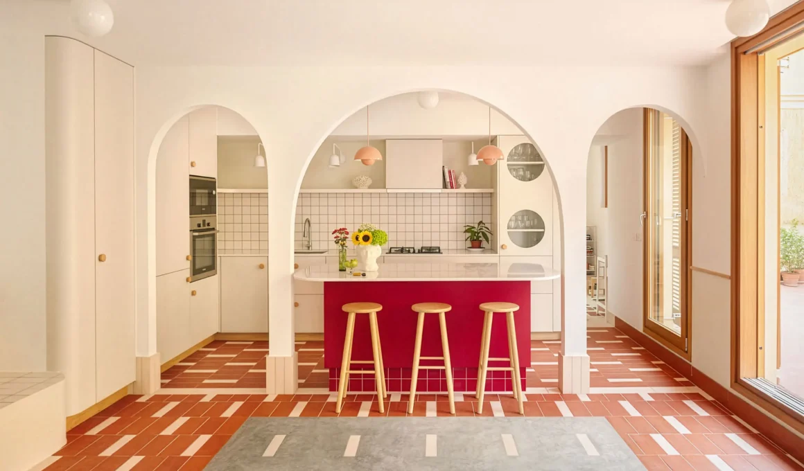 A vibrant kitchen island in burgundy red seen through three white architectural arches with terracotta tiled flooring.