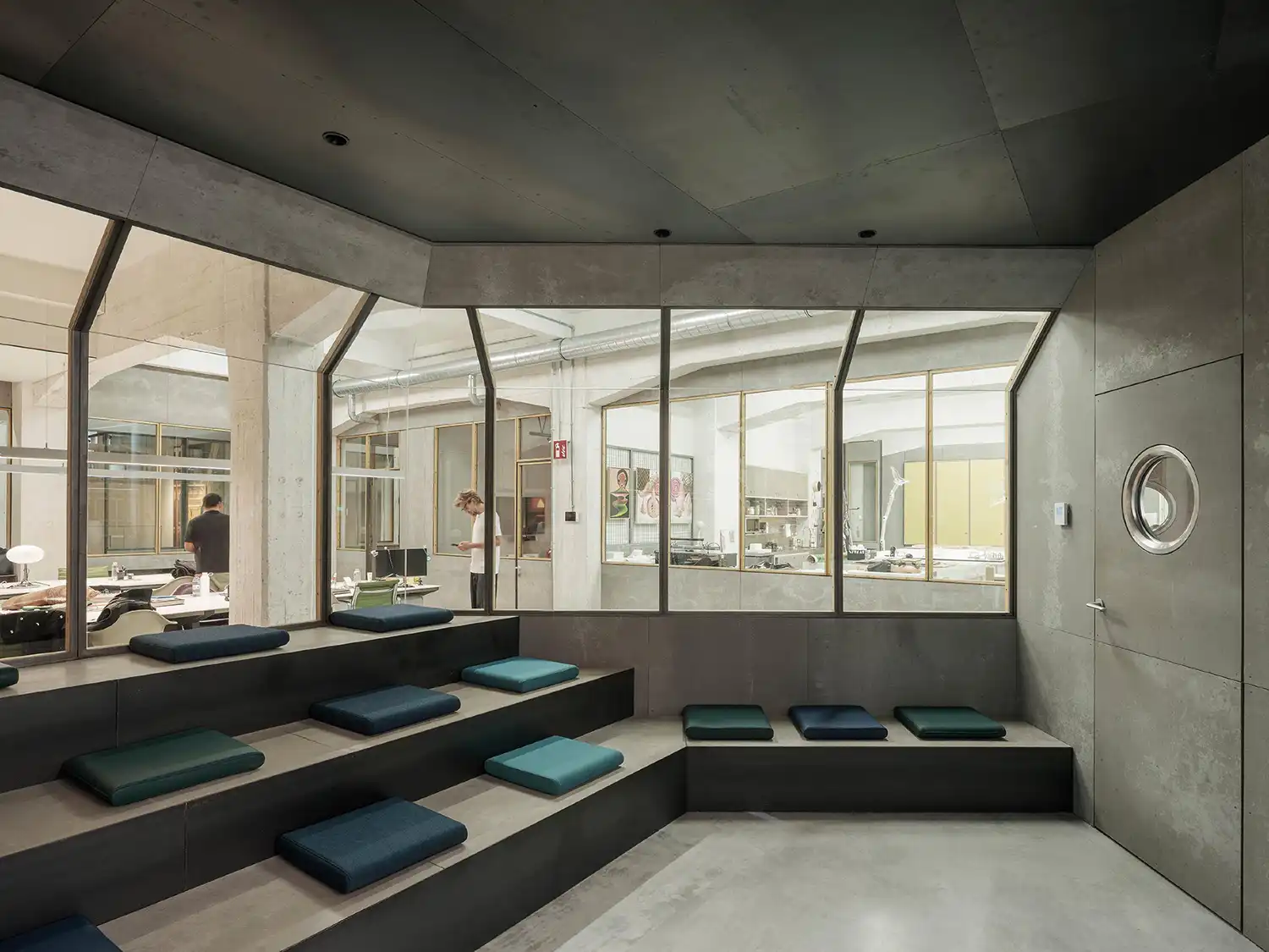 Tiered seating area made of dark recycled cement panels looking into a glazed workshop area.