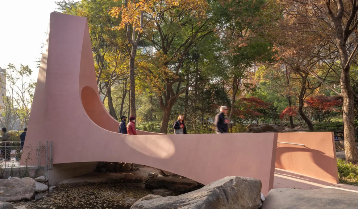 Monolithic pink concrete bridge by Hcch Studio spanning a stream in Huashan Greenland, Shanghai, surrounded by autumn trees and granite boulders.