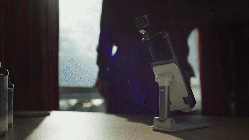 A smartphone mounted on a desk stand rotates its camera head to follow a person walking in the background.