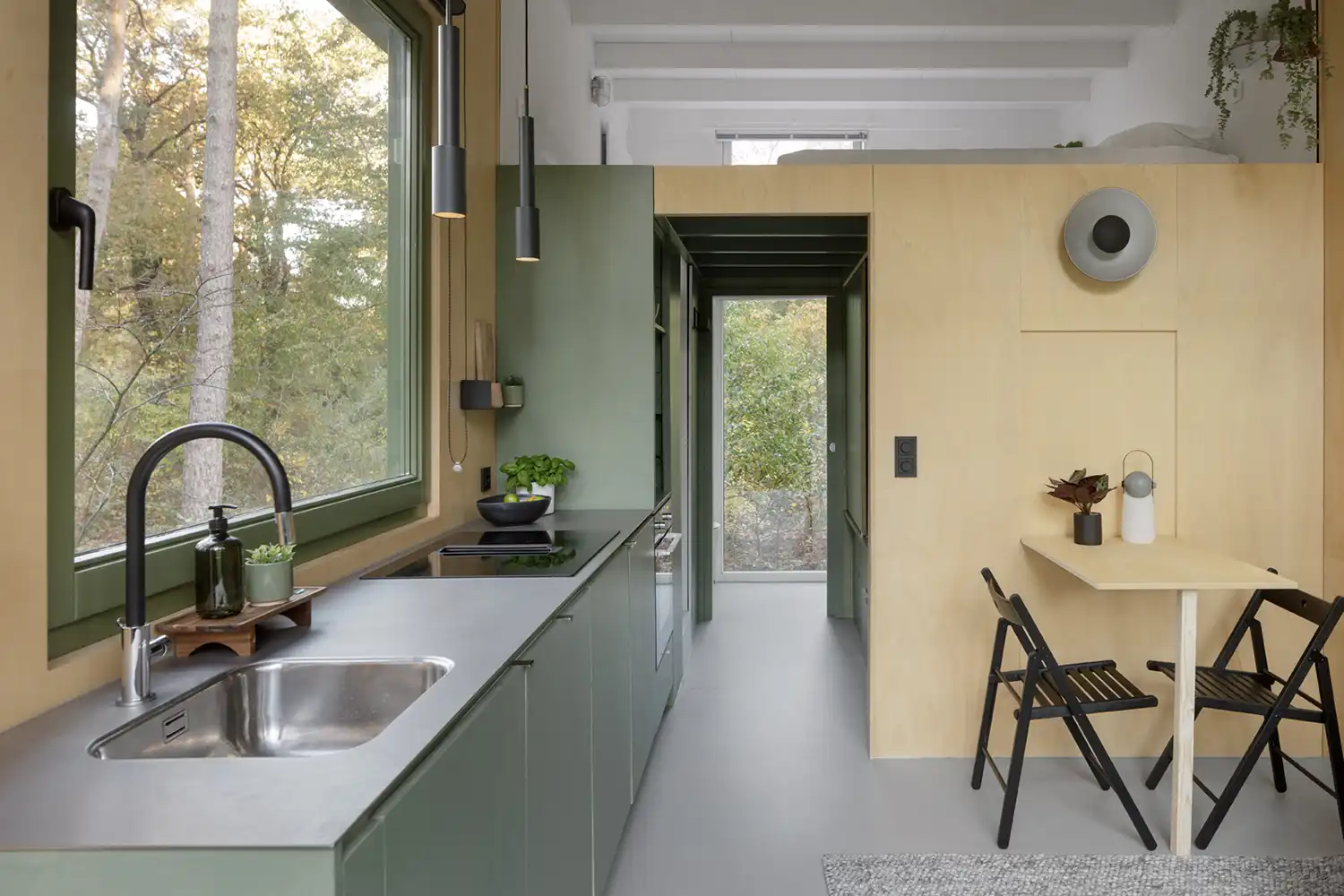 Interior view of a long, efficient green kitchen counter with a stainless steel sink and induction hob, positioned next to a large window facing the woods.