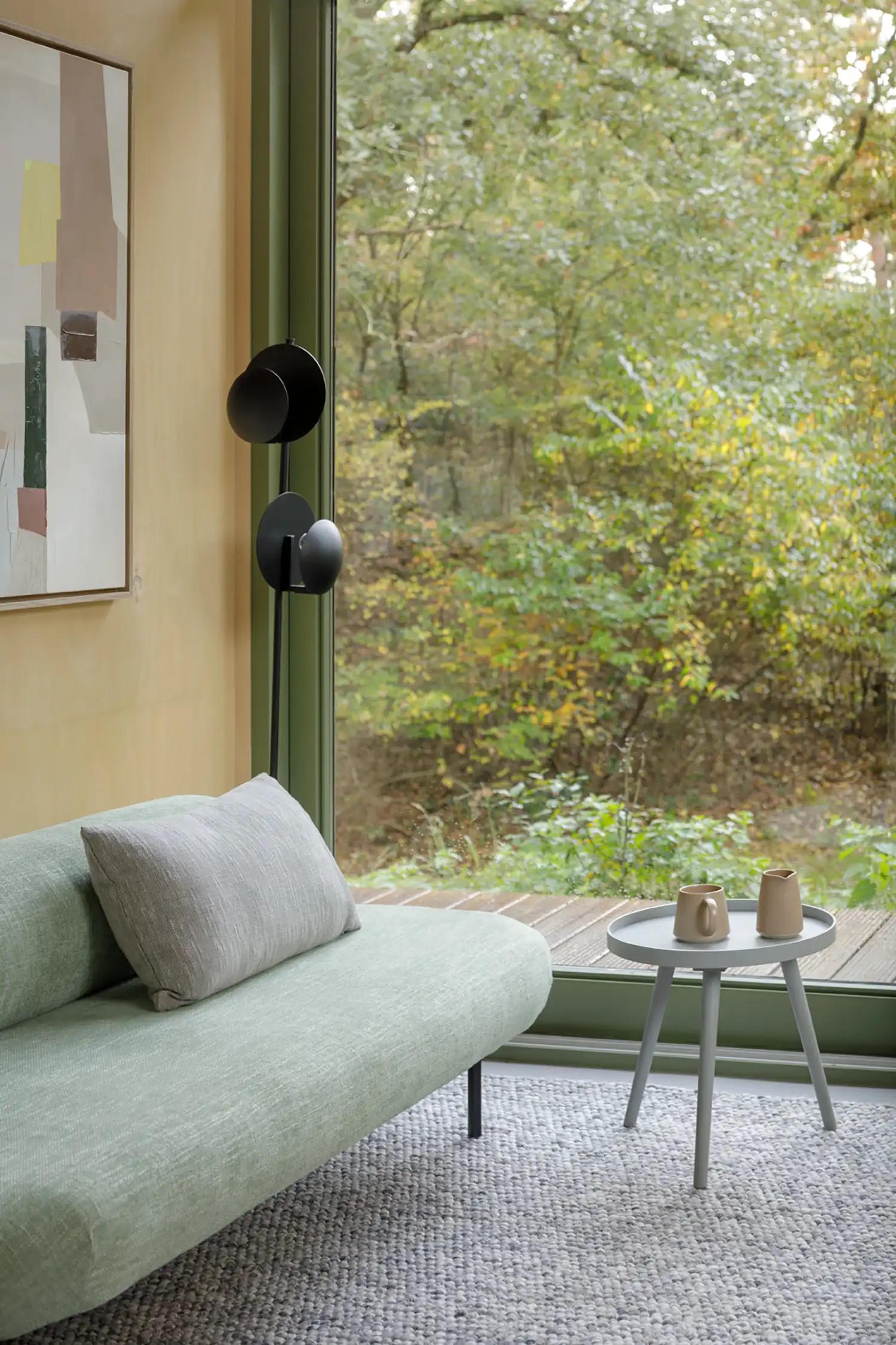 A light green sofa with a grey cushion sits on a woven grey rug next to a large floor-to-ceiling glass door, with a grey side table and cups.