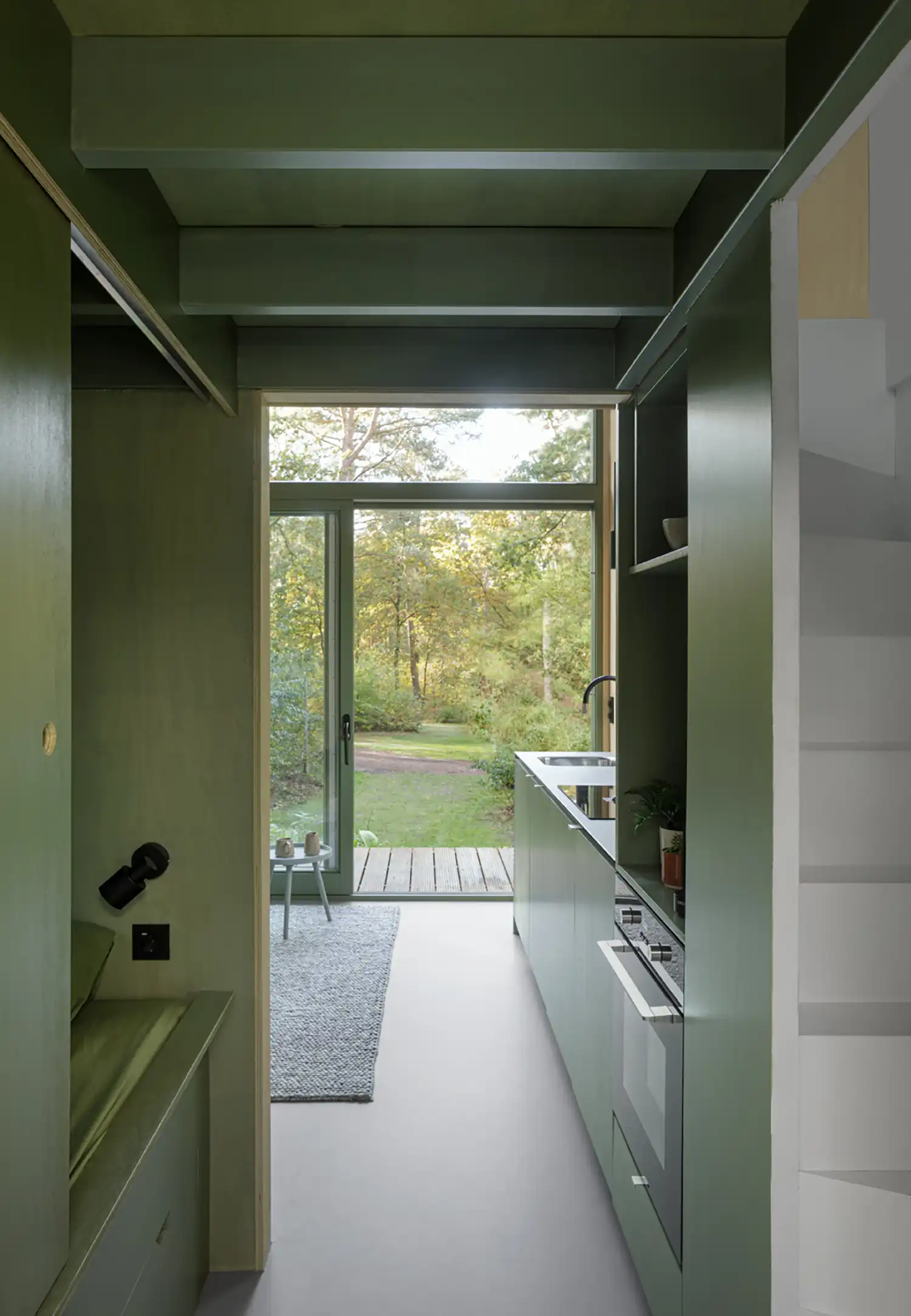 A view down the central axis of the cabin, looking out from the darker sleeping niche through the bright green kitchen corridor and out the glass door to the woods.