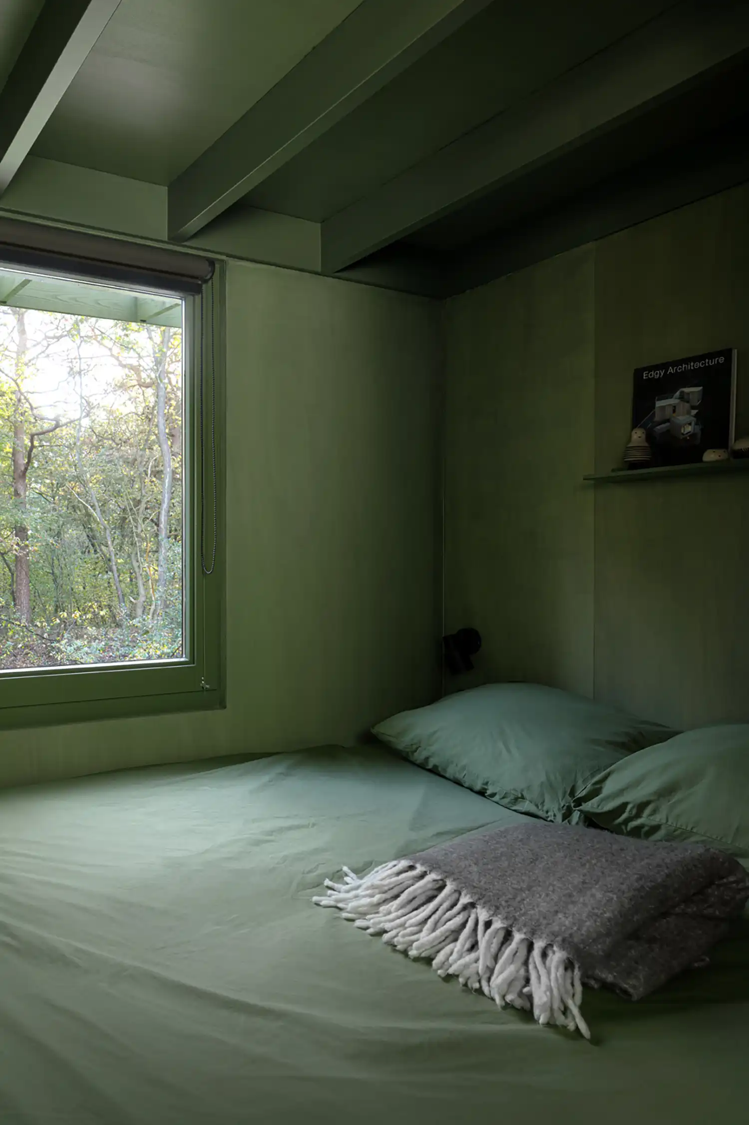 A close-up view of the minimalist sleeping niche, featuring a double bed with sage green bedding, a grey knitted blanket, a window, and a small shelf.
