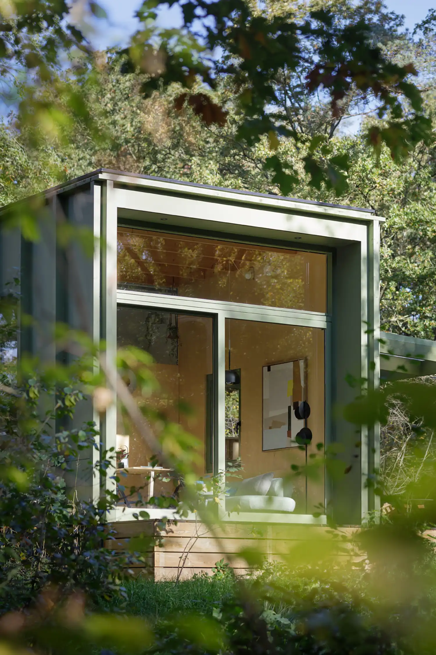 A close-up view through branches of the Lounge Lodge's glass facade, revealing the light wood interior and minimalist furnishings.