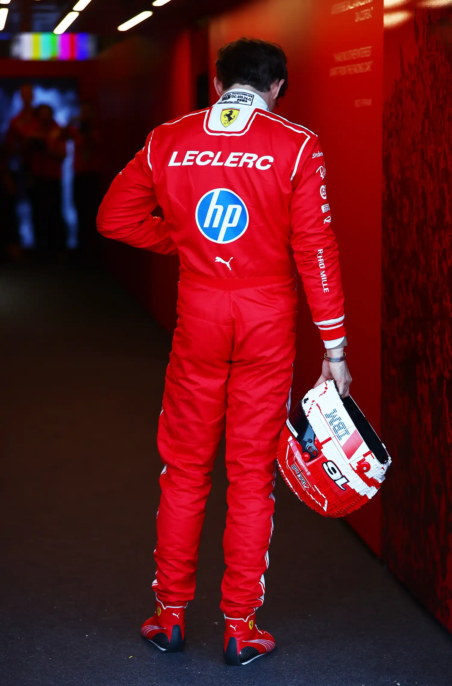 F1 driver Charles Leclerc from behind in a red racing suit carrying a life-sized LEGO helmet.