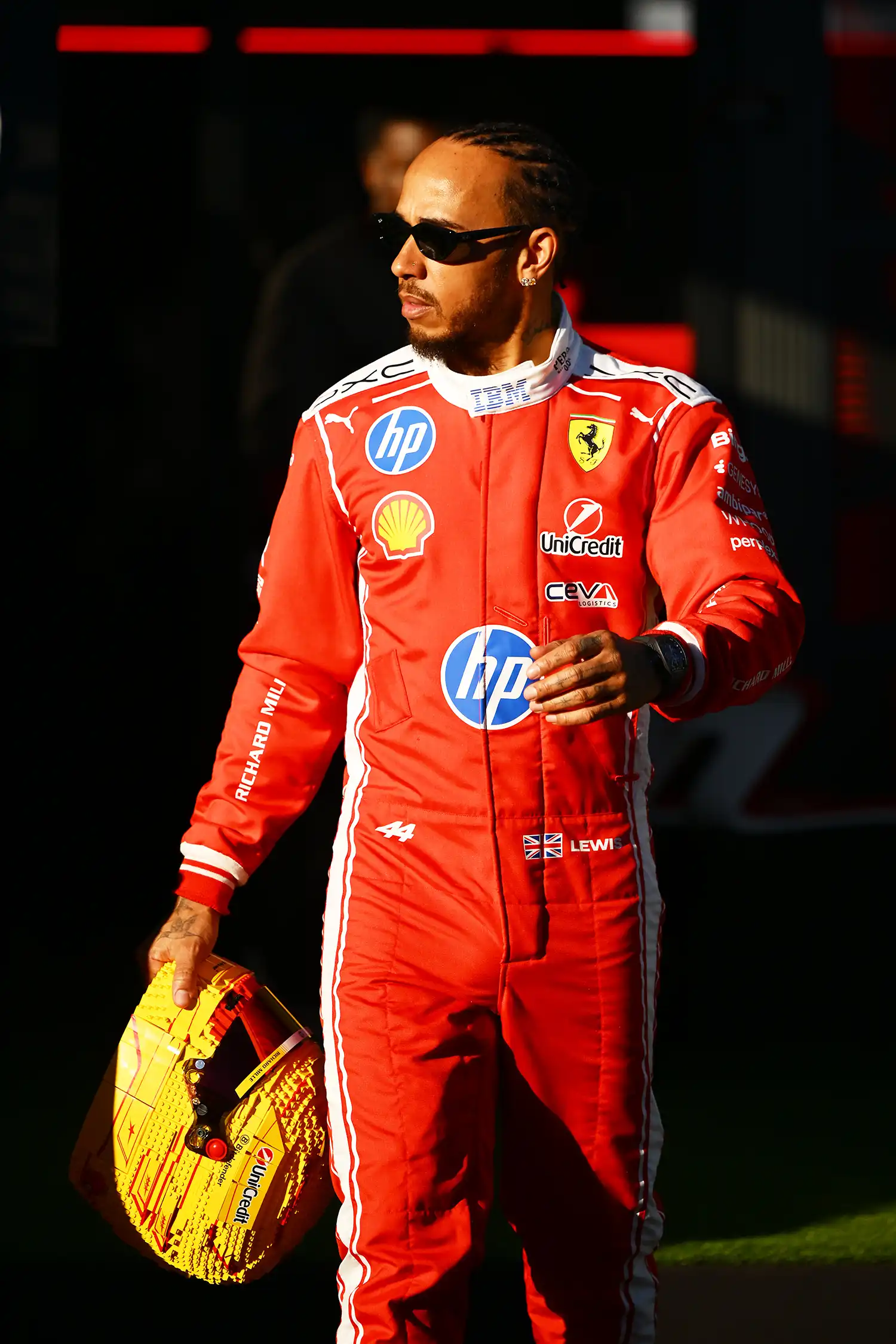 Lewis Hamilton in a Scuderia Ferrari HP racing suit walking with a yellow LEGO helmet.