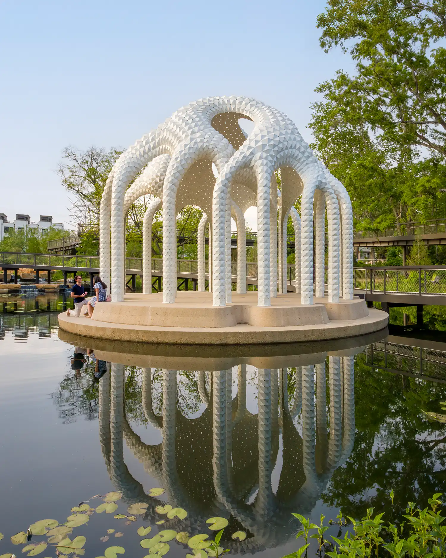 Ground level view of L’île Folie pavilion reflecting in the pond water under a clear sky.