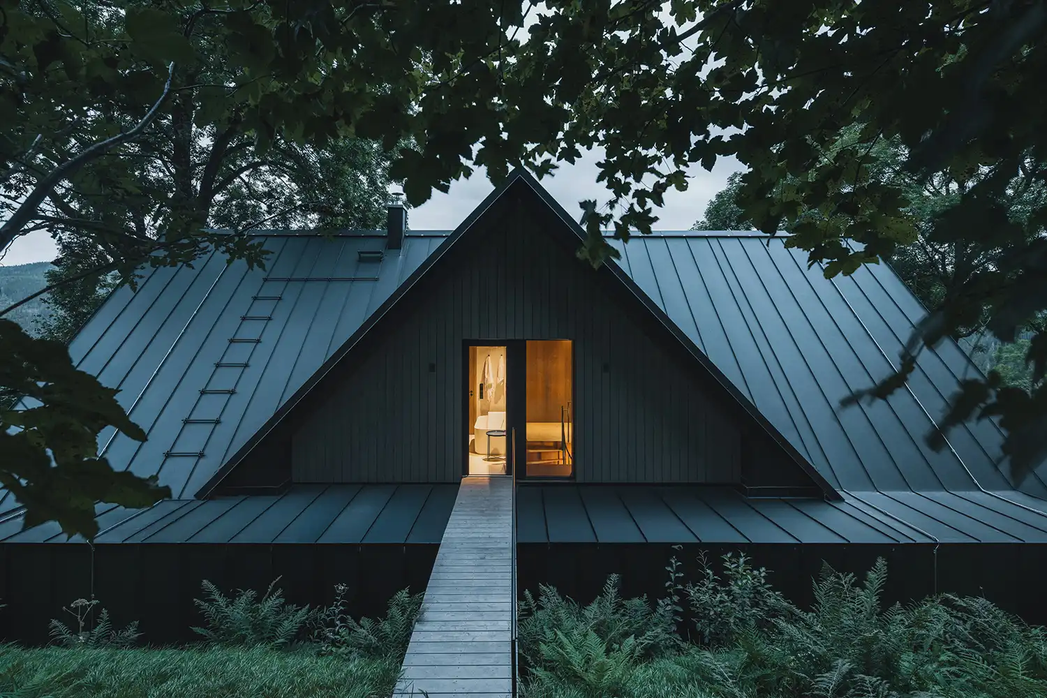 Evening view of the cottage's rear entrance with a wooden bridge leading to a lit interior.