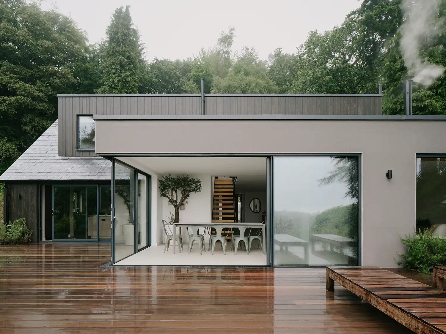 A wide exterior shot of the rear facade of Beech Cottage, featuring a wet timber deck and open sliding glass doors leading into the dining space.