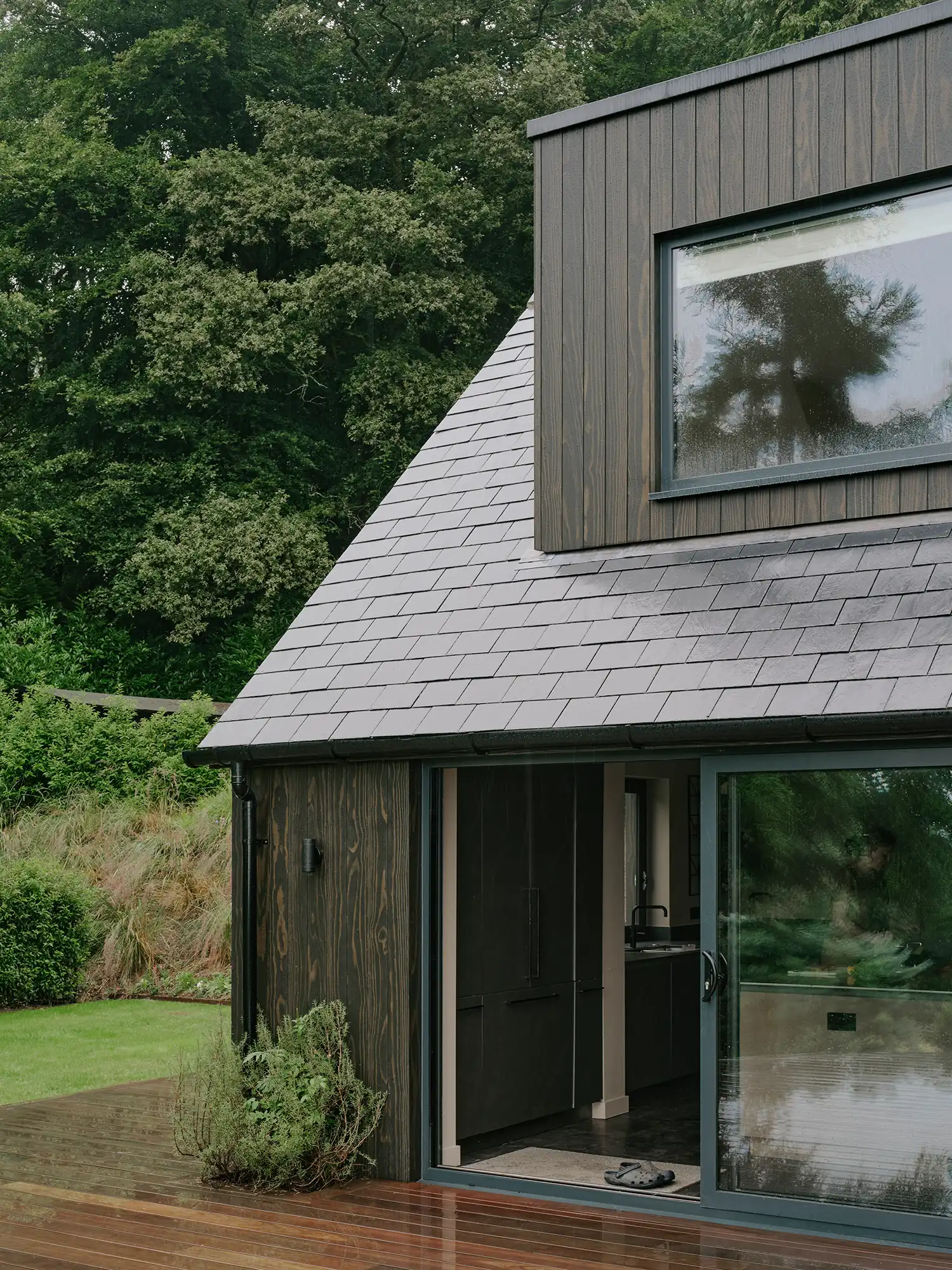 Detail shot of the cottage's exterior, showing the intersection of dark-stained timber cladding, slate roof tiles, and a sliding glass door.