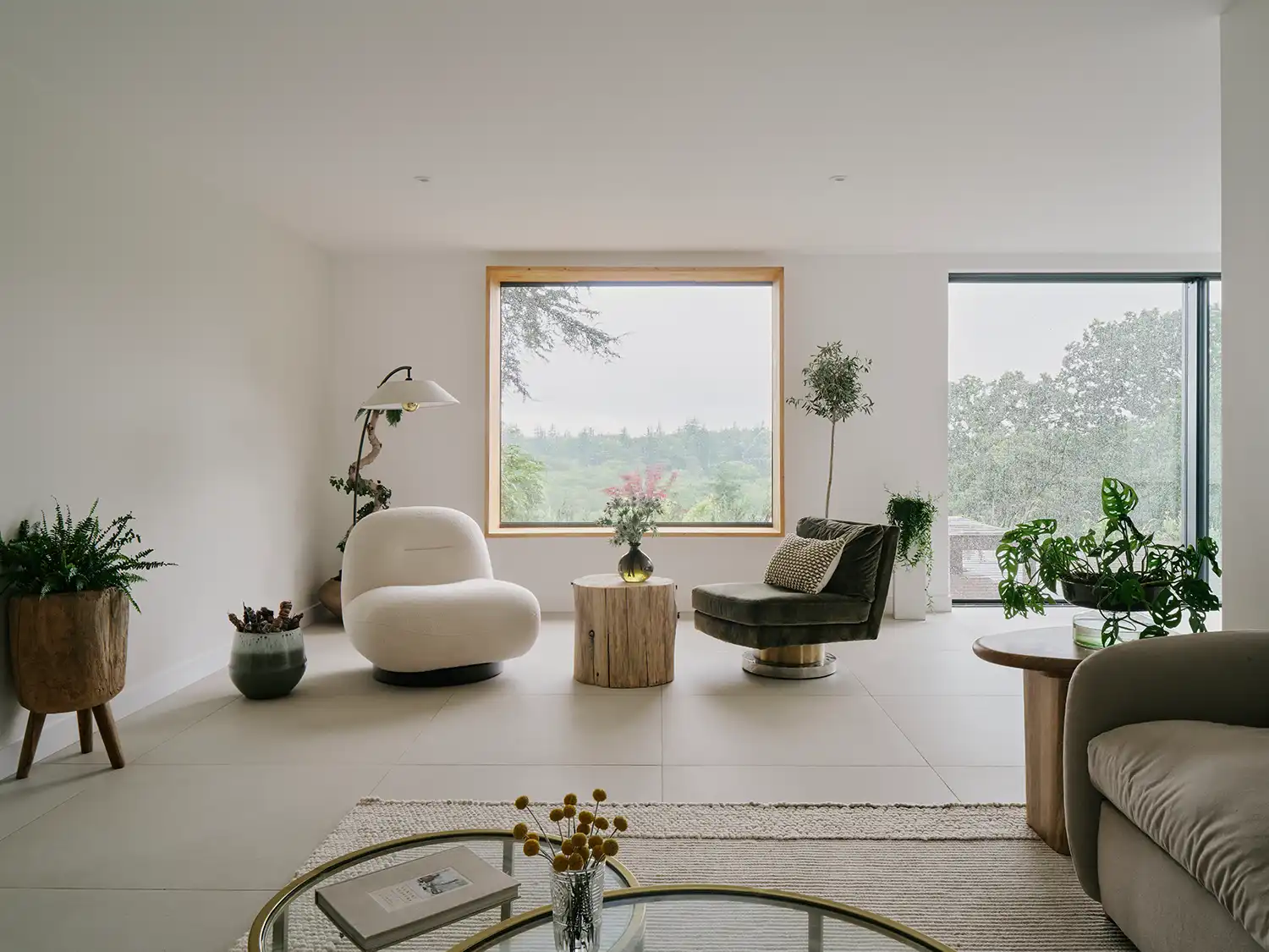A bright, minimalist living room with white furniture, large-format tiles, and a large square picture window framing the forest.
