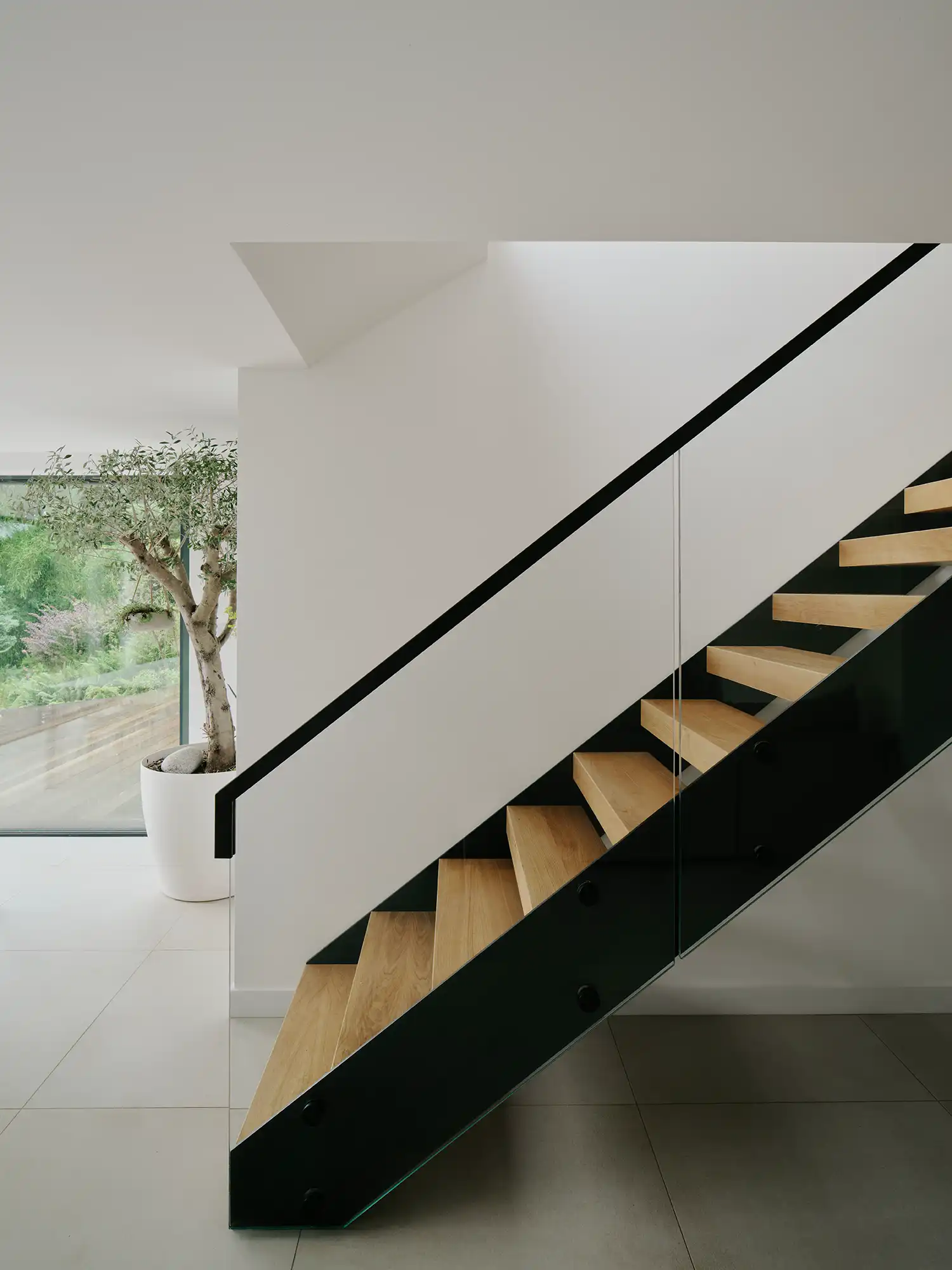 Interior view of an open-tread timber staircase with a glass balustrade and black metal stringer, set against a white wall.