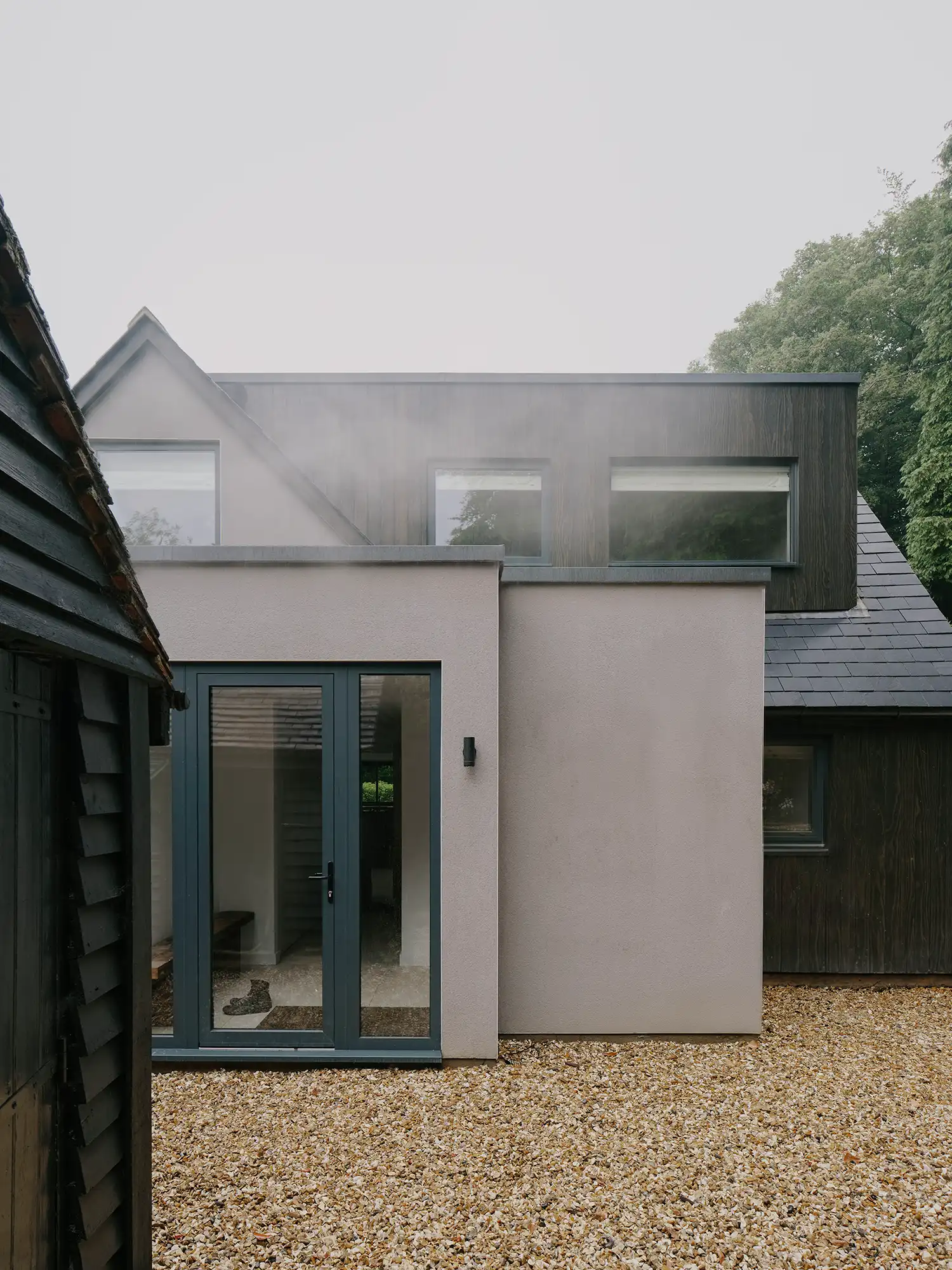 Exterior shot of the entrance area with a gravel driveway and the contrast of a dark timber shed against the new rendered volumes.