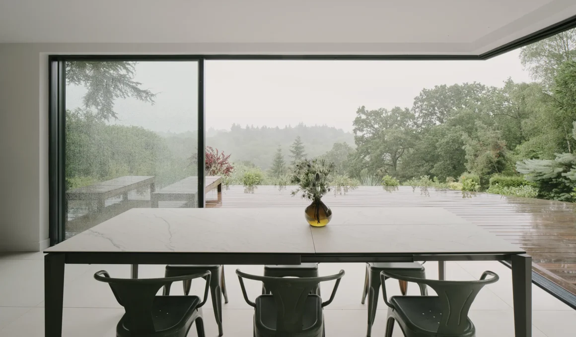 Interior view of a minimalist dining area with a long table, green chairs, and a large corner glass window overlooking a misty Surrey Hills landscape.