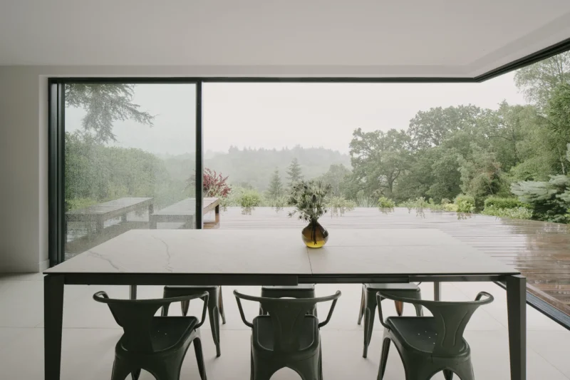 Interior view of a minimalist dining area with a long table, green chairs, and a large corner glass window overlooking a misty Surrey Hills landscape.