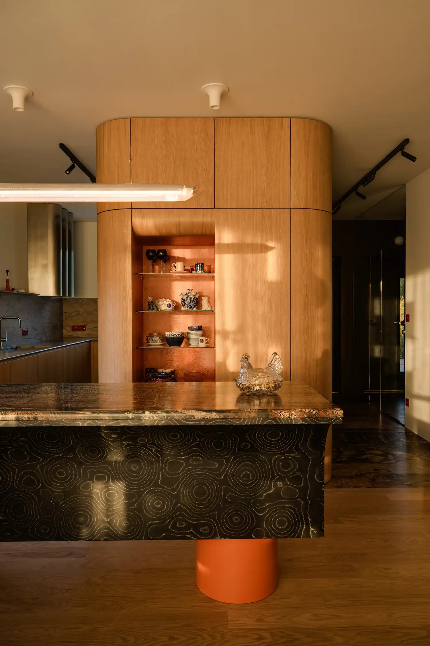 Sculptural kitchen island with orange base and patterned veneer next to a rounded oak volume.