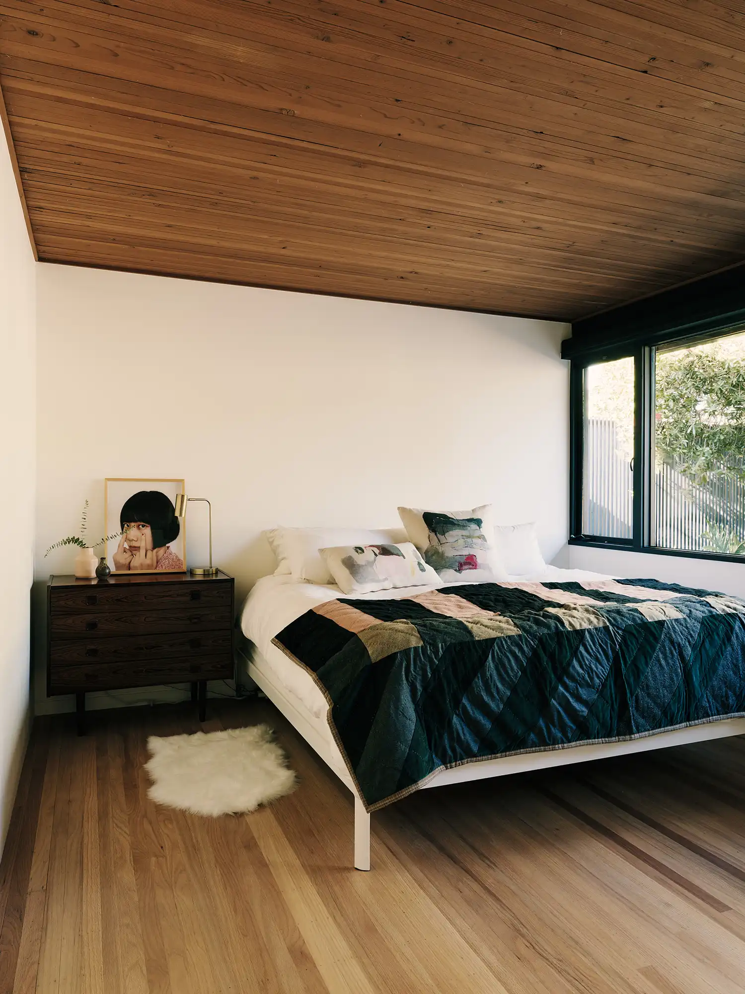 Minimalist bedroom with mahogany ceiling, white walls, and a large window overlooking trees.