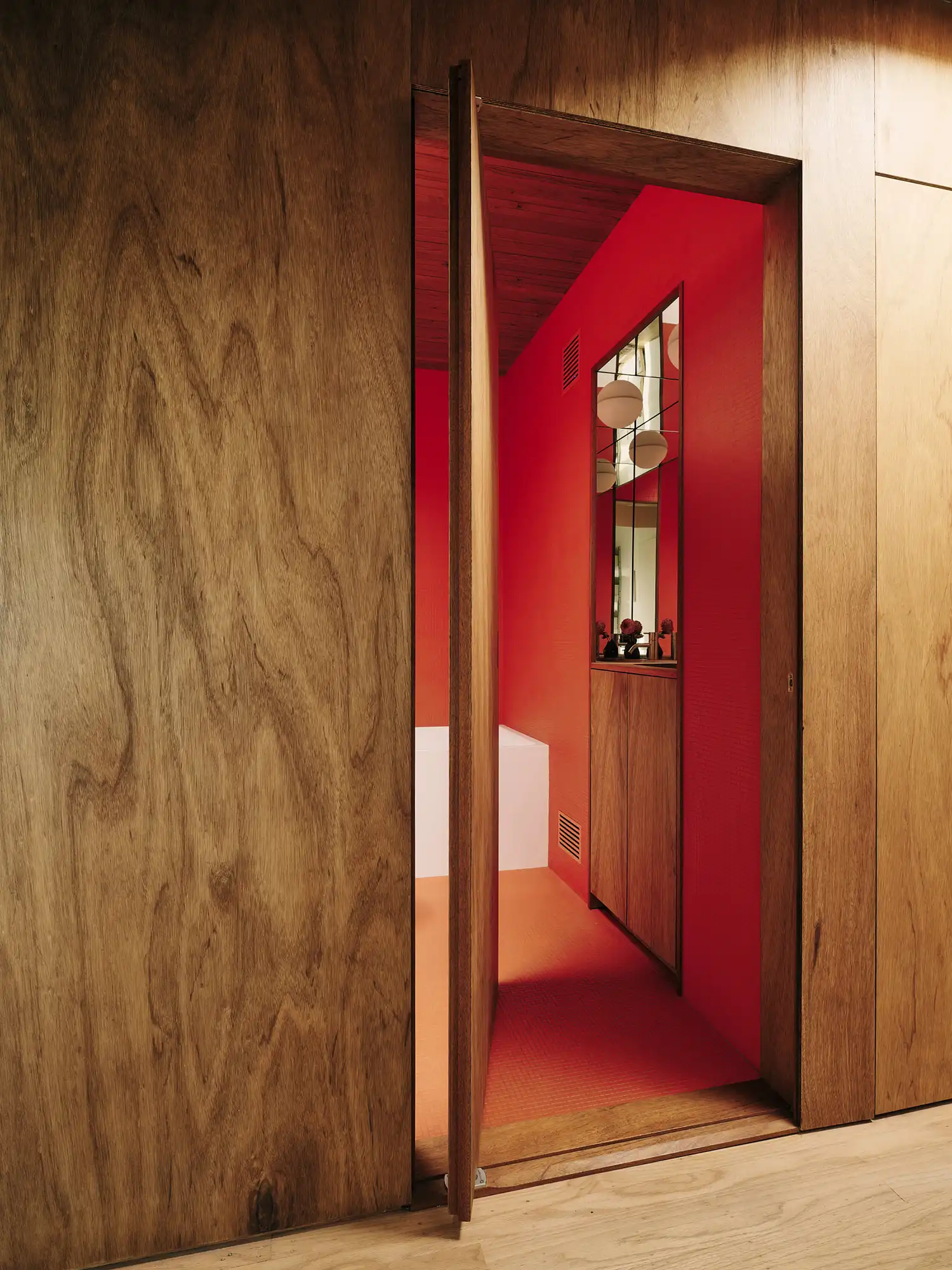 An open wood-veneered door revealing a bright red tiled bathroom interior.