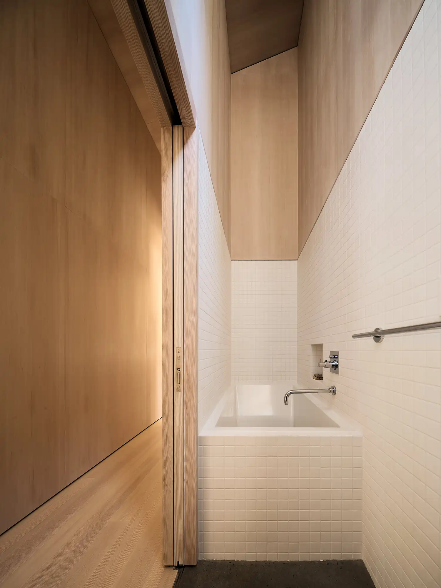 Narrow bathroom with white square tiles and a deep white soaking tub set against warm wood-paneled walls.