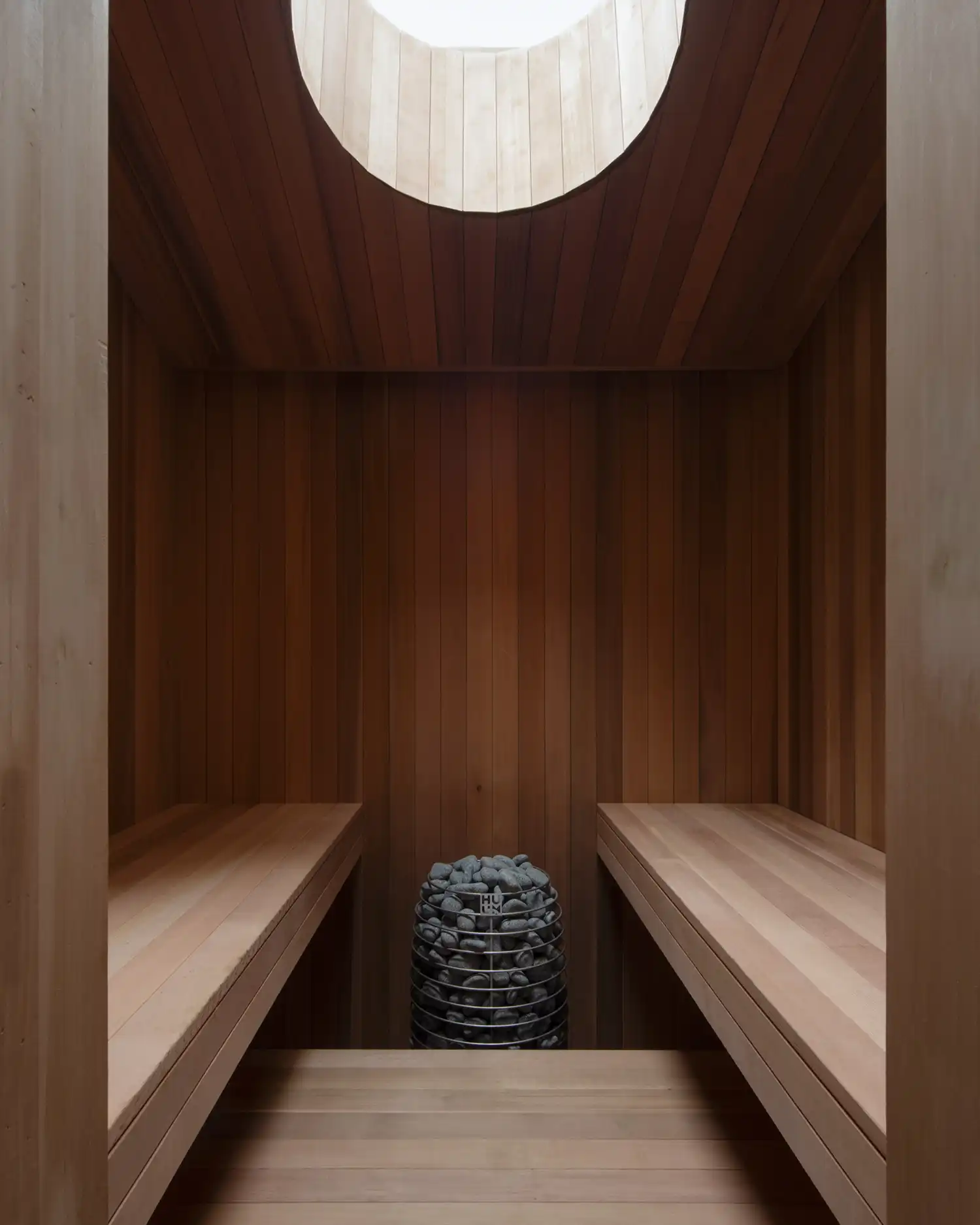 Minimalist wood-lined sauna with a central stone heater and circular oculus.