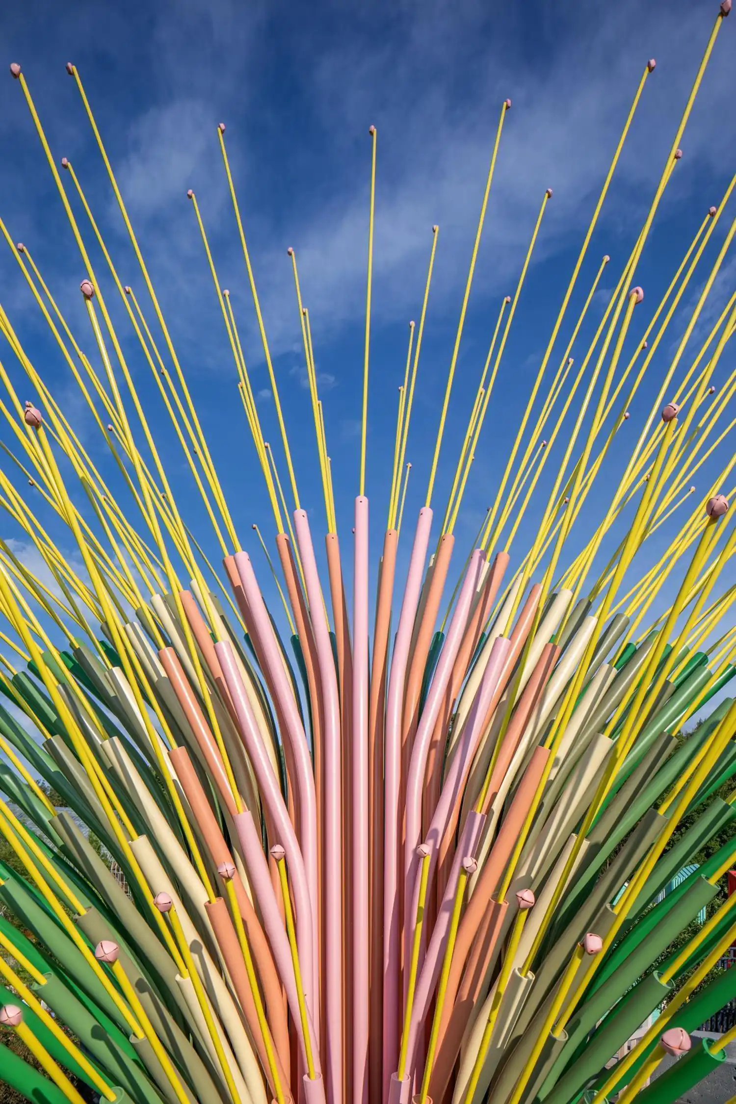 Vertical close-up of a cluster of steel tubes in shades of pink, tan, and green topped with yellow flexible rods.