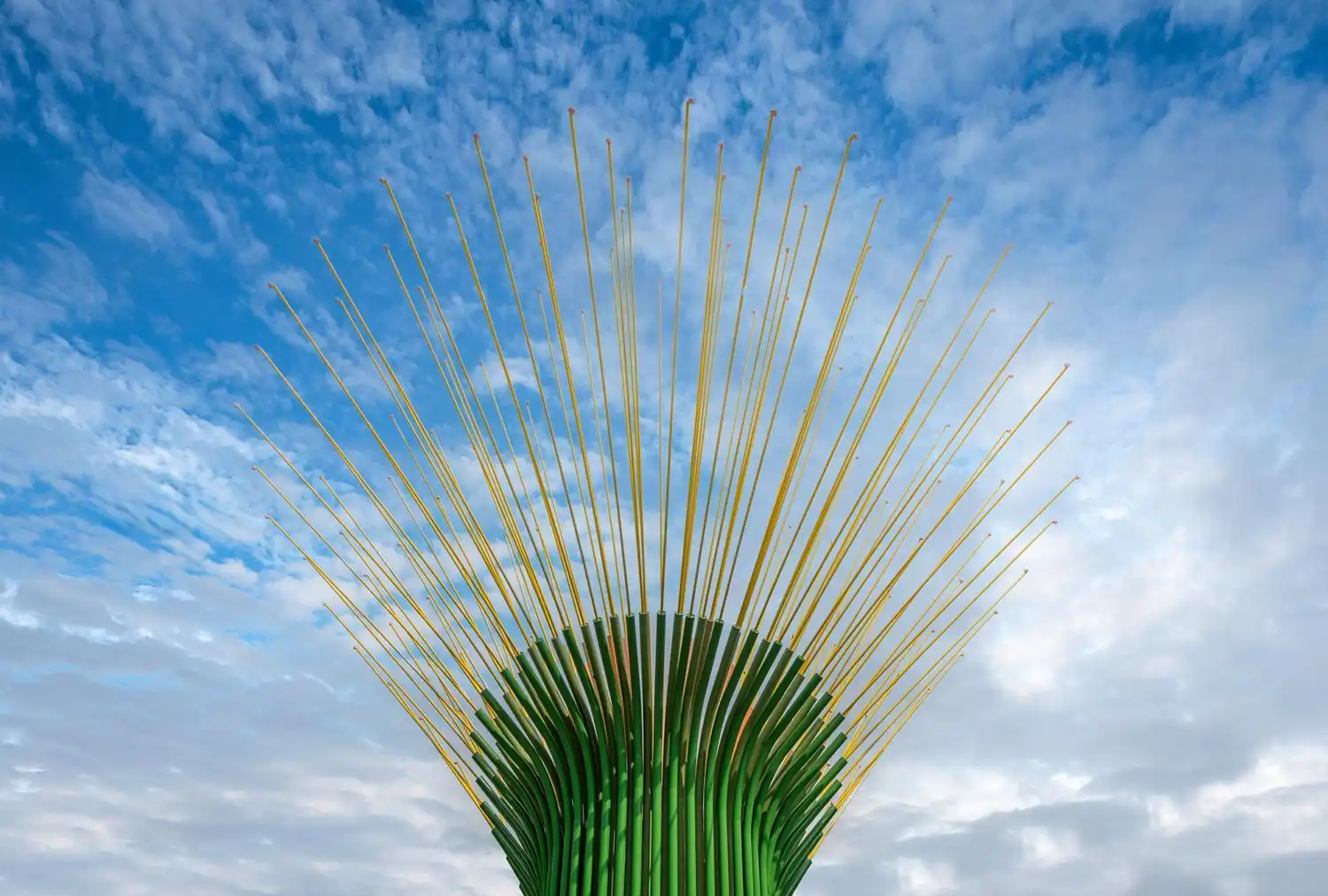 Wide shot of the top of the installation showing dozens of thin yellow rods tipped with small bells fanning out against a cloudy sky.