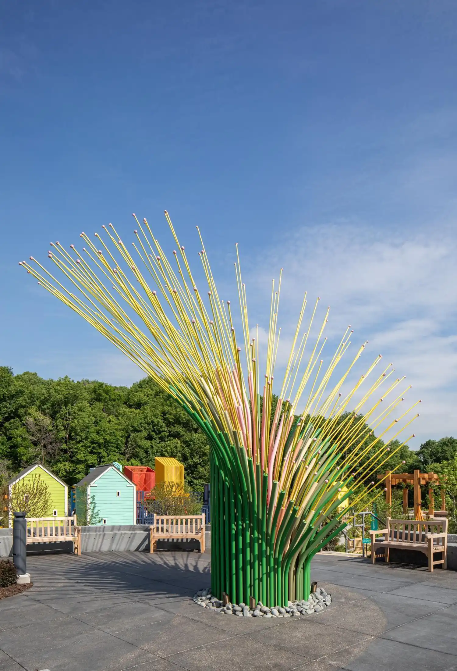 Side view of the leaning sculpture in a park with colorful small playhouses and wooden benches in the background.