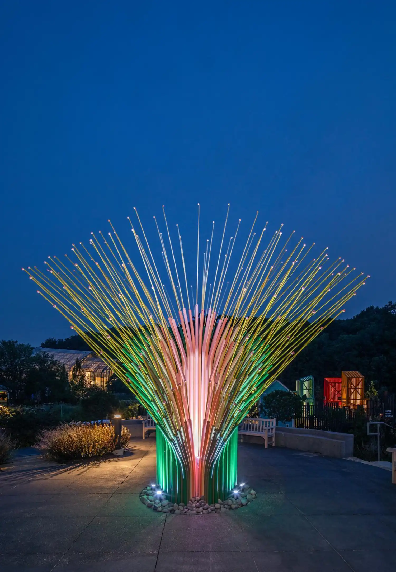 The kinetic sculpture glowing with internal pink and green light at dusk in a dark garden.