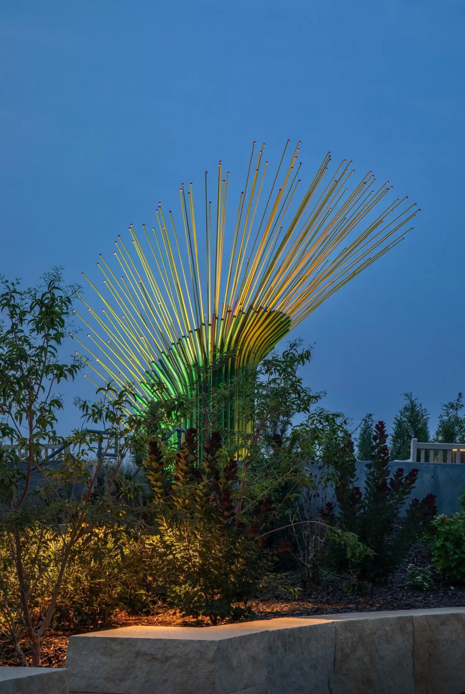 Nocturnal profile of the sculpture illuminated in green behind dense foreground vegetation.
